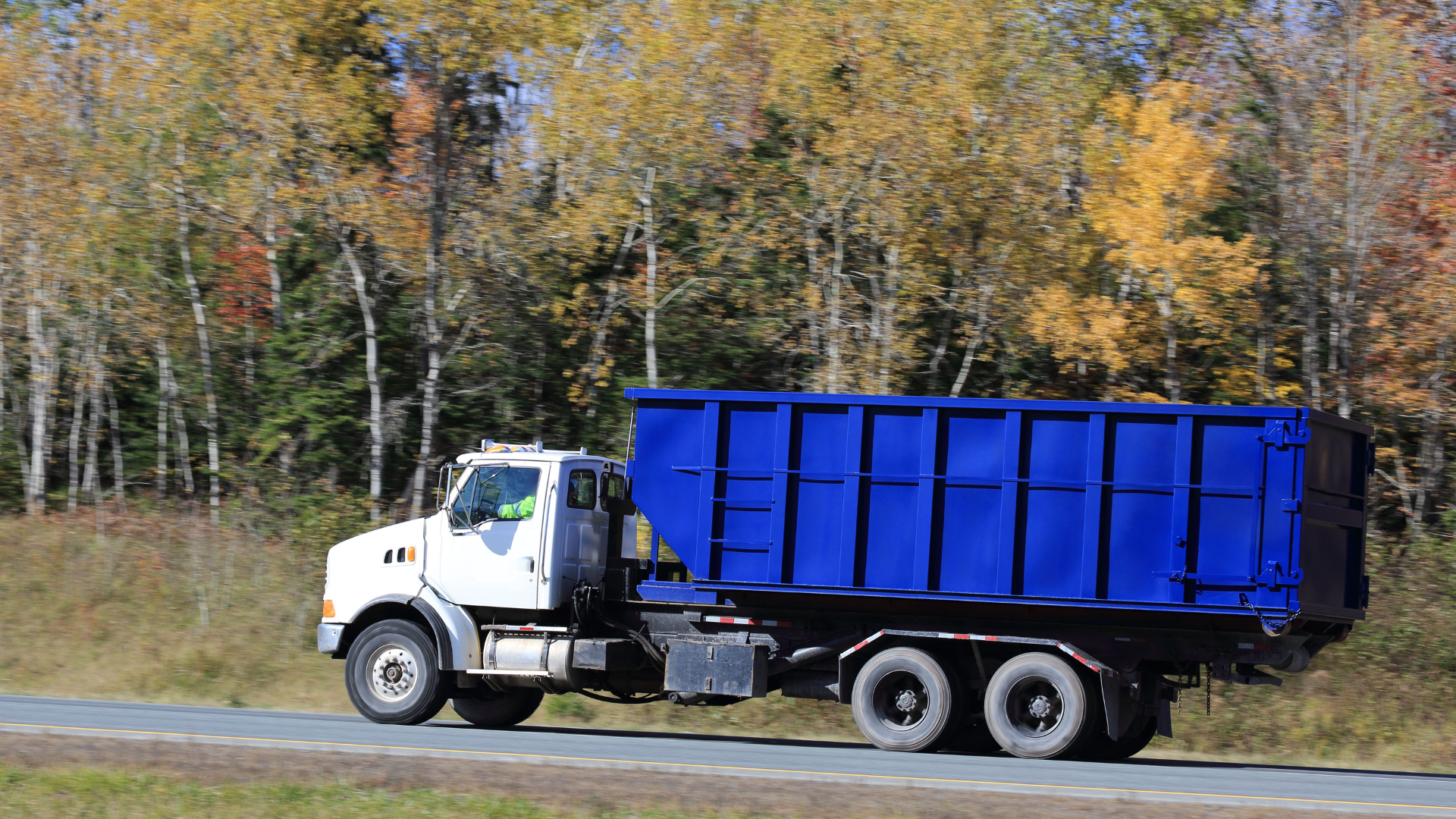 White dump truck with a blue bed driving on a road beside autumn trees