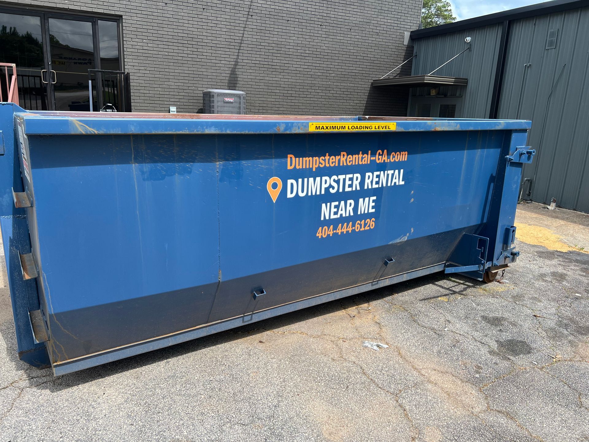 Two people smiling in front of a dumpster rental sign reading “Need a Dumpster?”
