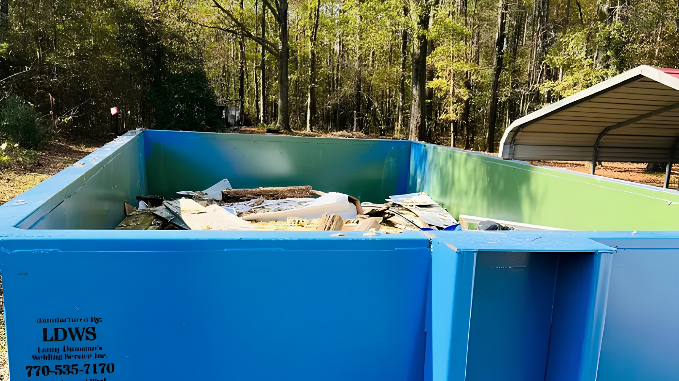 Blue and green dumpster filled with debris outdoors near trees and a shed
