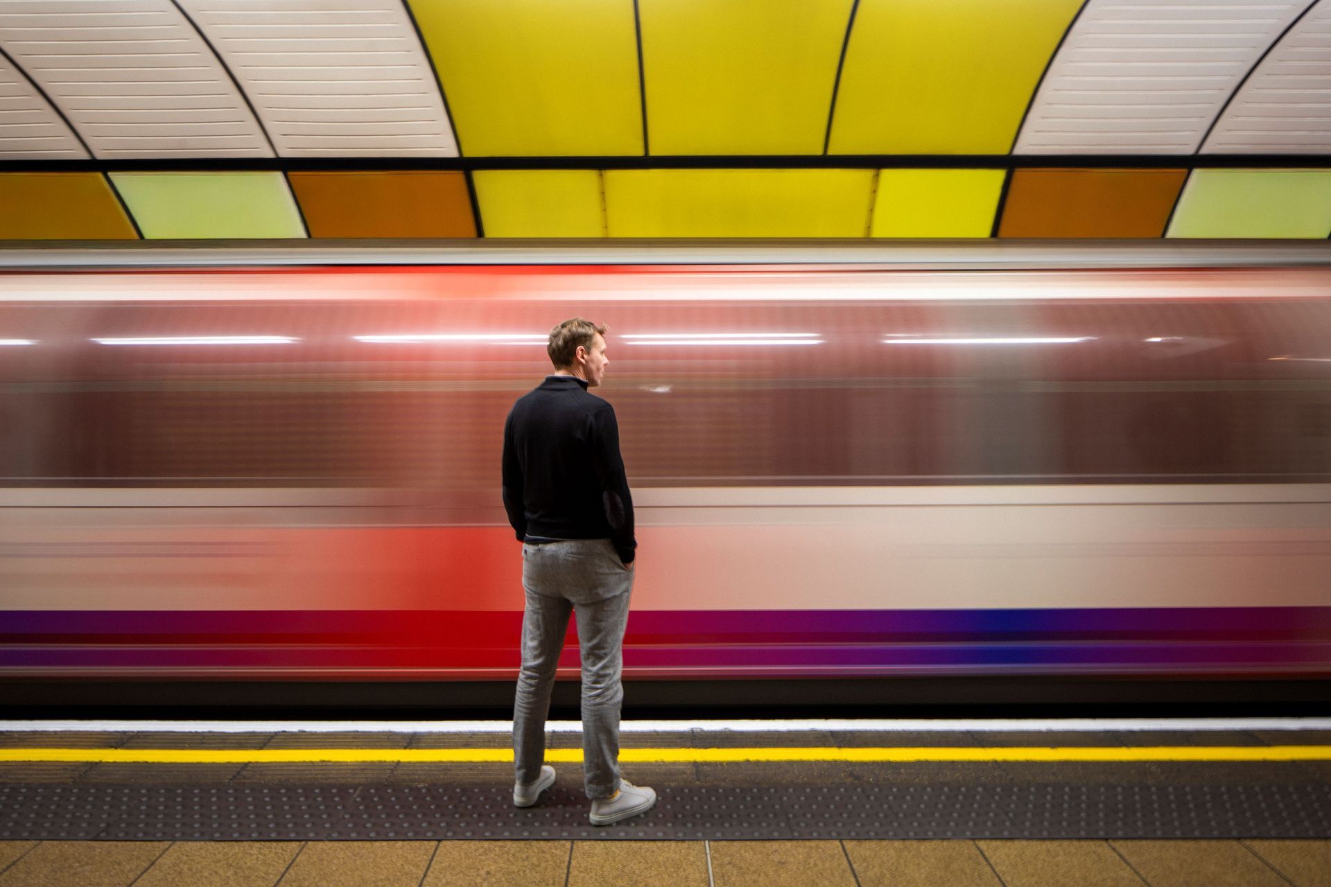 Person standing on a subway platform as a train rushes by, with yellow and red station accents.