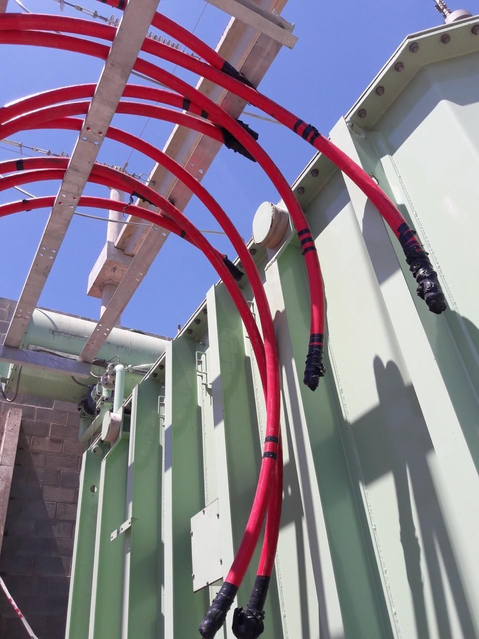 Cables eléctricos rojos conectados a un gran transformador verde al aire libre contra un cielo azul.
