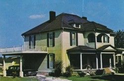 Two-story light funeral home with a covered porch and car port, under a blue sky.