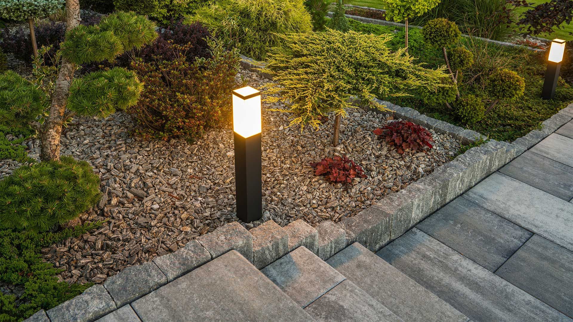 A lamp post is lit up in a garden next to a set of stairs.