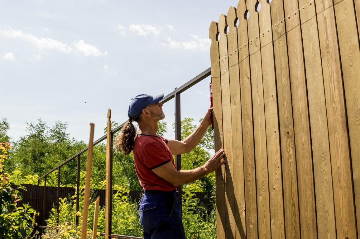 Person in red shirt and blue cap fixing a wooden fence outdoors on a sunny day.