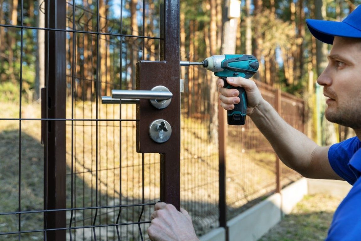 Man in blue uniform uses a power drill to install a gate lock on a brown metal fence.