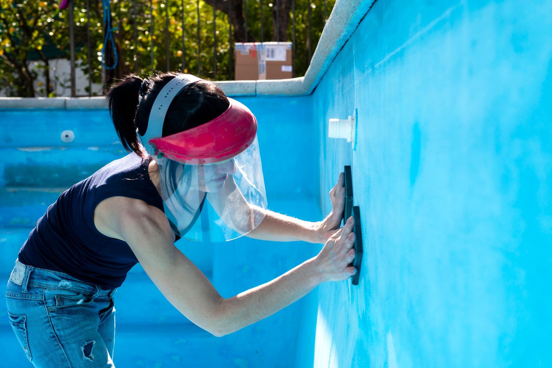 Woman wearing face shield sanding pool wall during residential pool repair and surface maintenance.