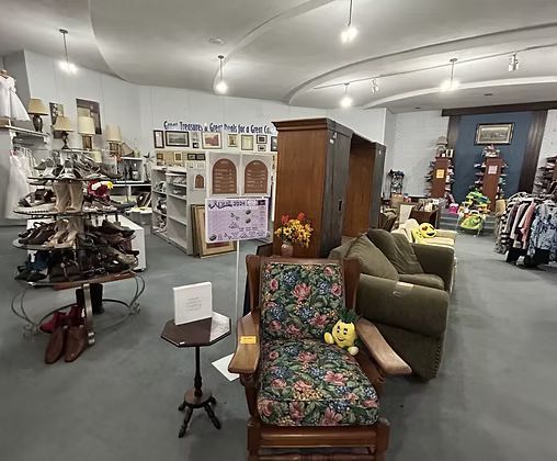 Thrift store interior with furniture, shoes, and miscellaneous items on display under bright lighting.