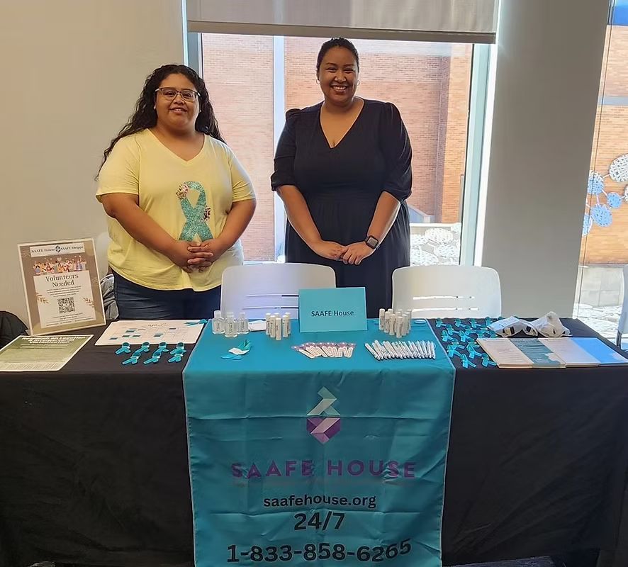 Two people stand behind a table with teal branding. The table has brochures and ribbon. They are in a room with a window.