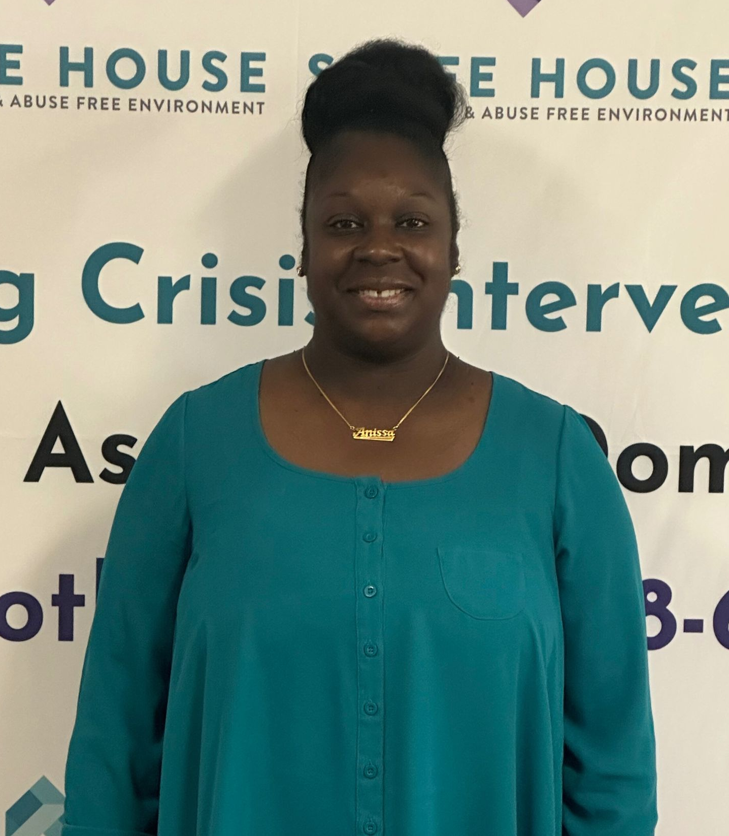 Woman in teal top smiles in front of a sign for SAAFE HOUSE crisis intervention services.