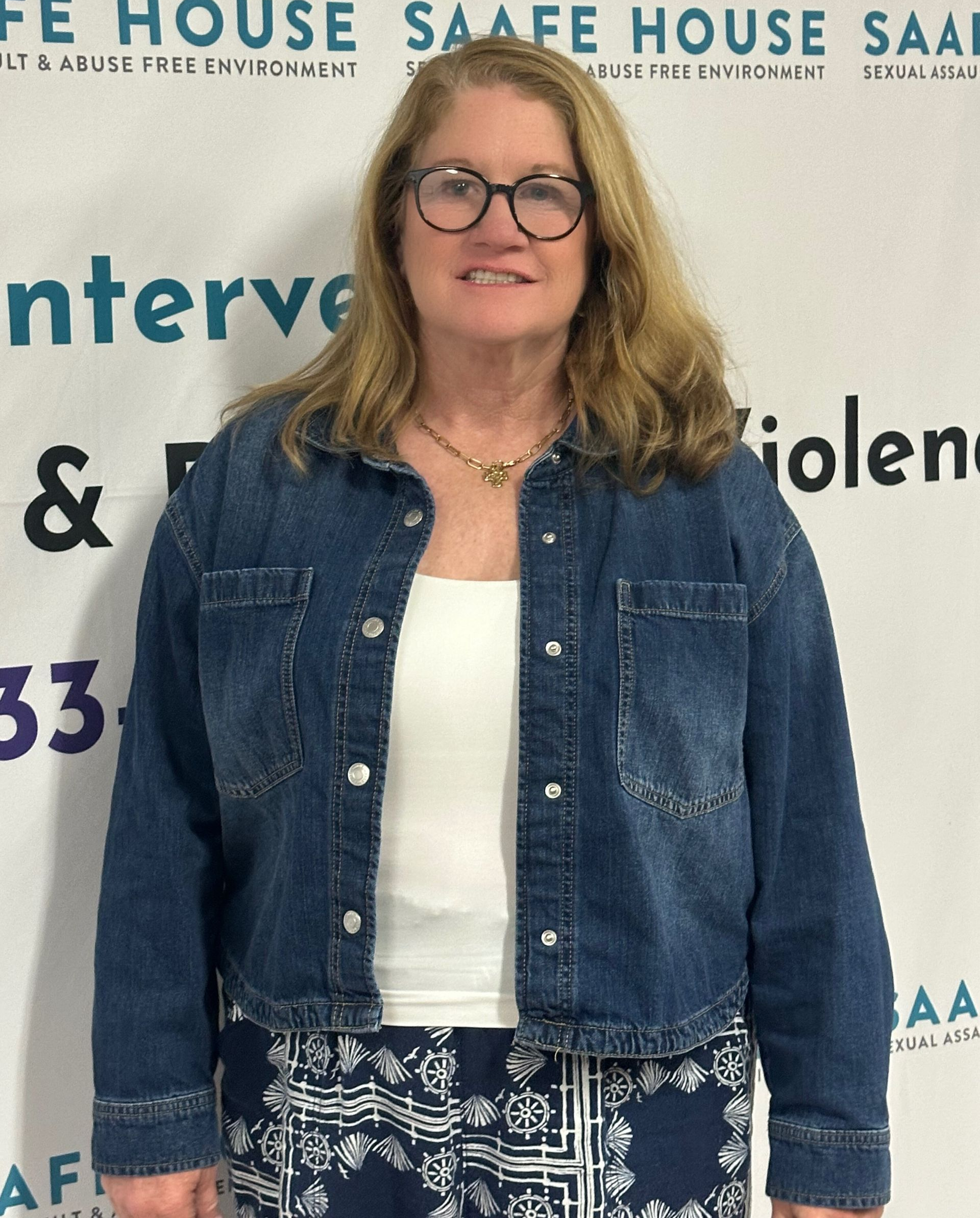 Woman in denim jacket, white top, patterned skirt, glasses. Standing in front of a banner with text.