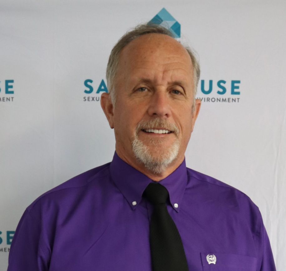 Man with graying hair and goatee wearing a purple shirt and black tie, smiling, in front of a banner with text.