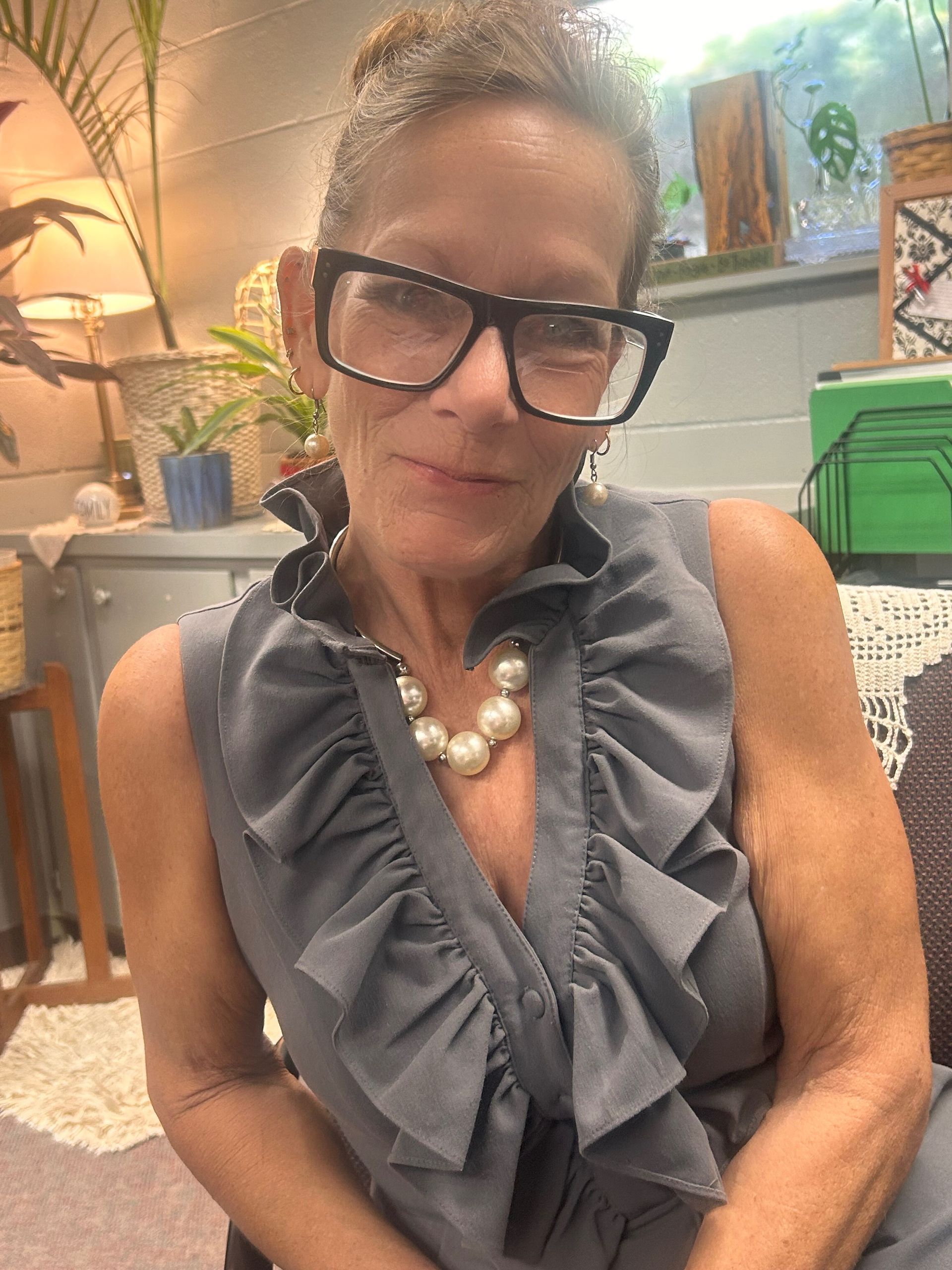 Woman wearing glasses and pearl necklace, in a gray top with ruffles, indoors.