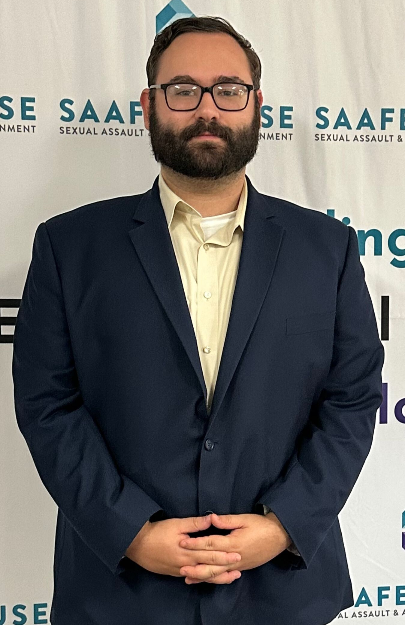 Man with glasses and beard wearing a navy blazer over a light yellow button-down shirt