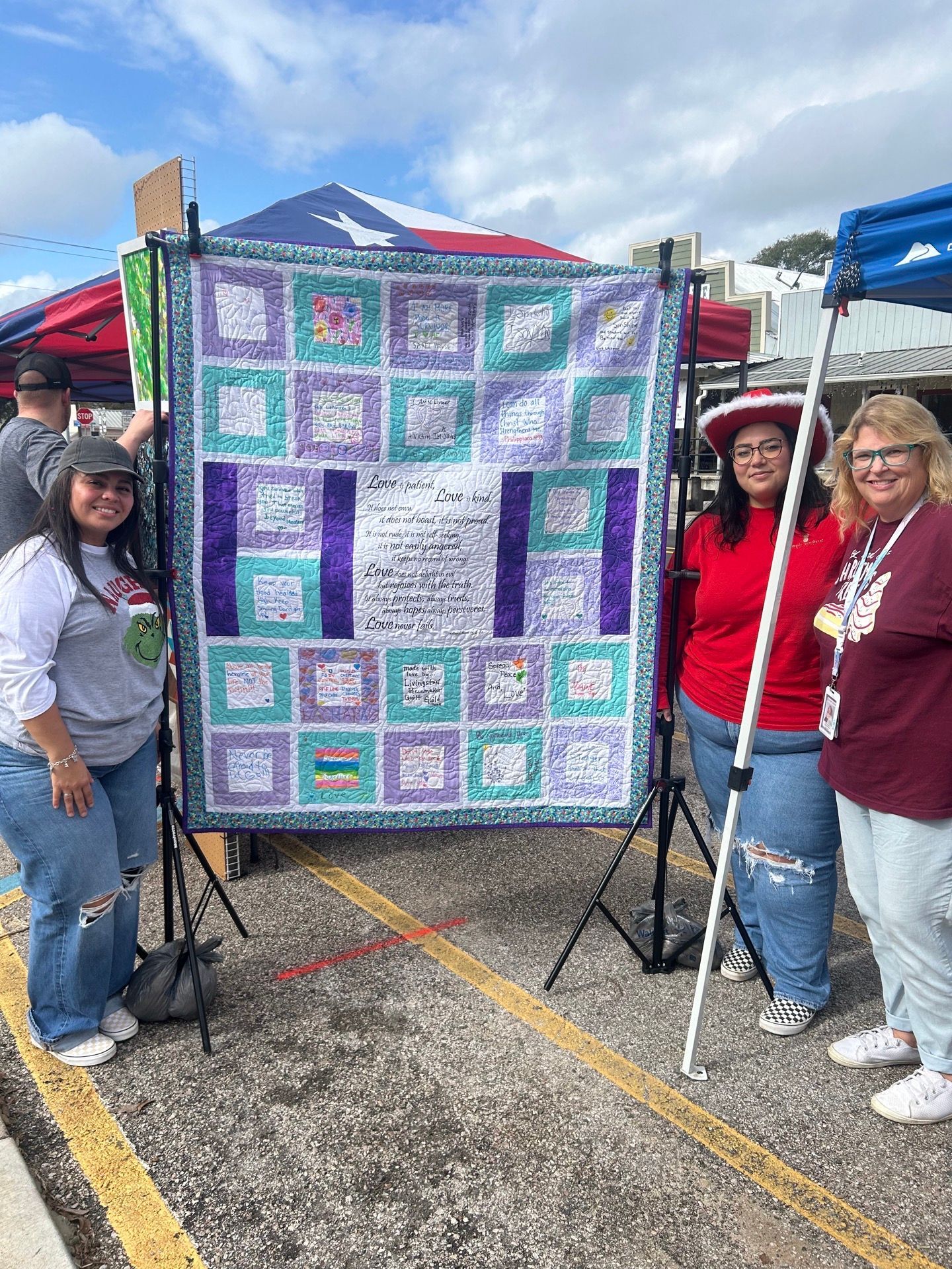 Three people stand by a quilt, smiling. The quilt is displayed outdoors on a stand. Purple and turquoise squares, and white text are visible.