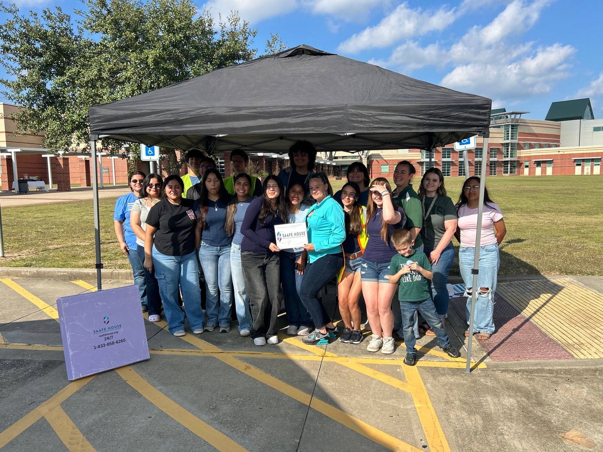 Group of people under a canopy, holding a sign in a parking lot. Buildings and a grassy area in the background.