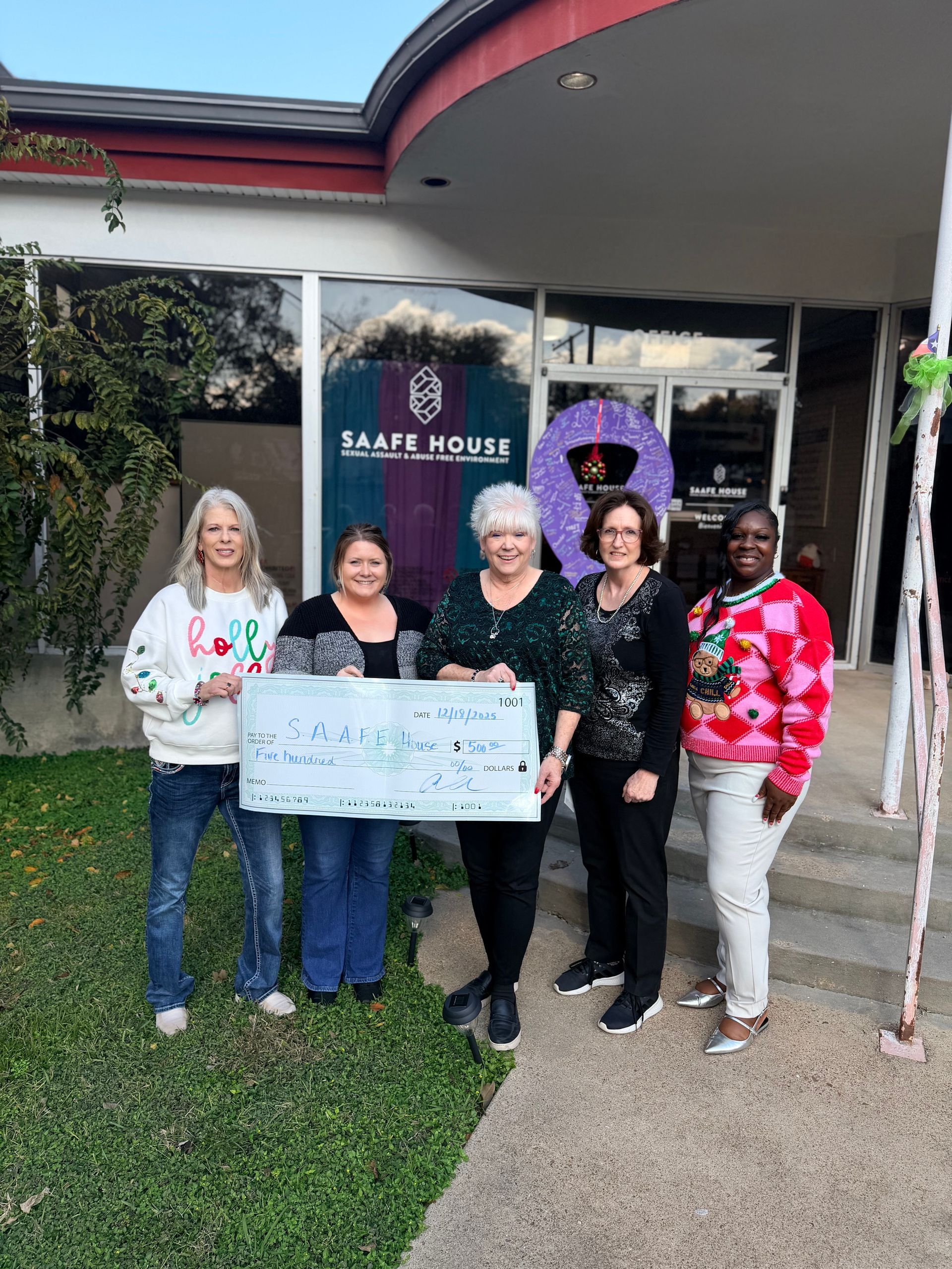 Five women stand outside a building, holding a large check. Building has a purple wreath.