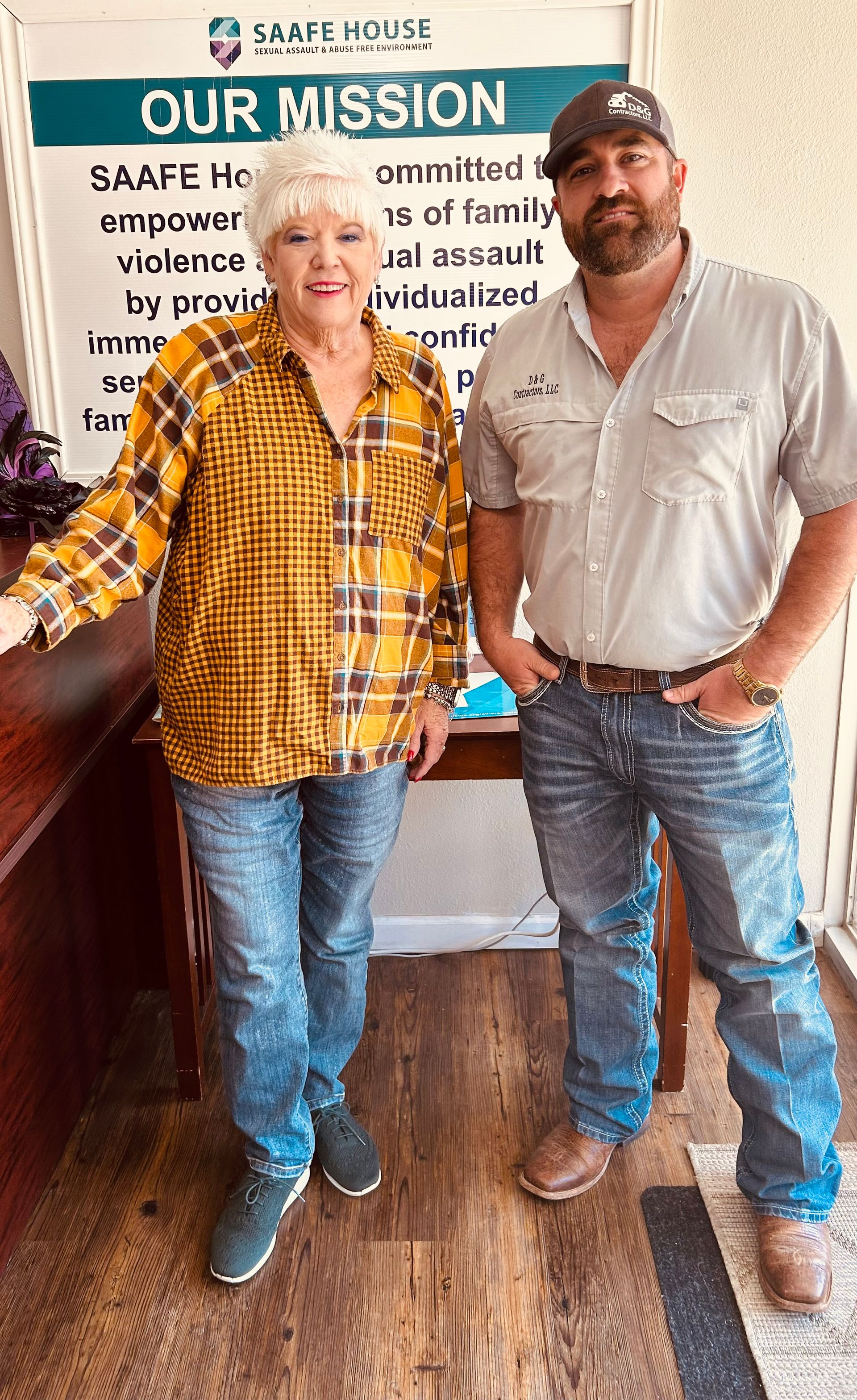 Woman and man standing near a sign. The woman wears a yellow plaid shirt, the man a gray shirt.