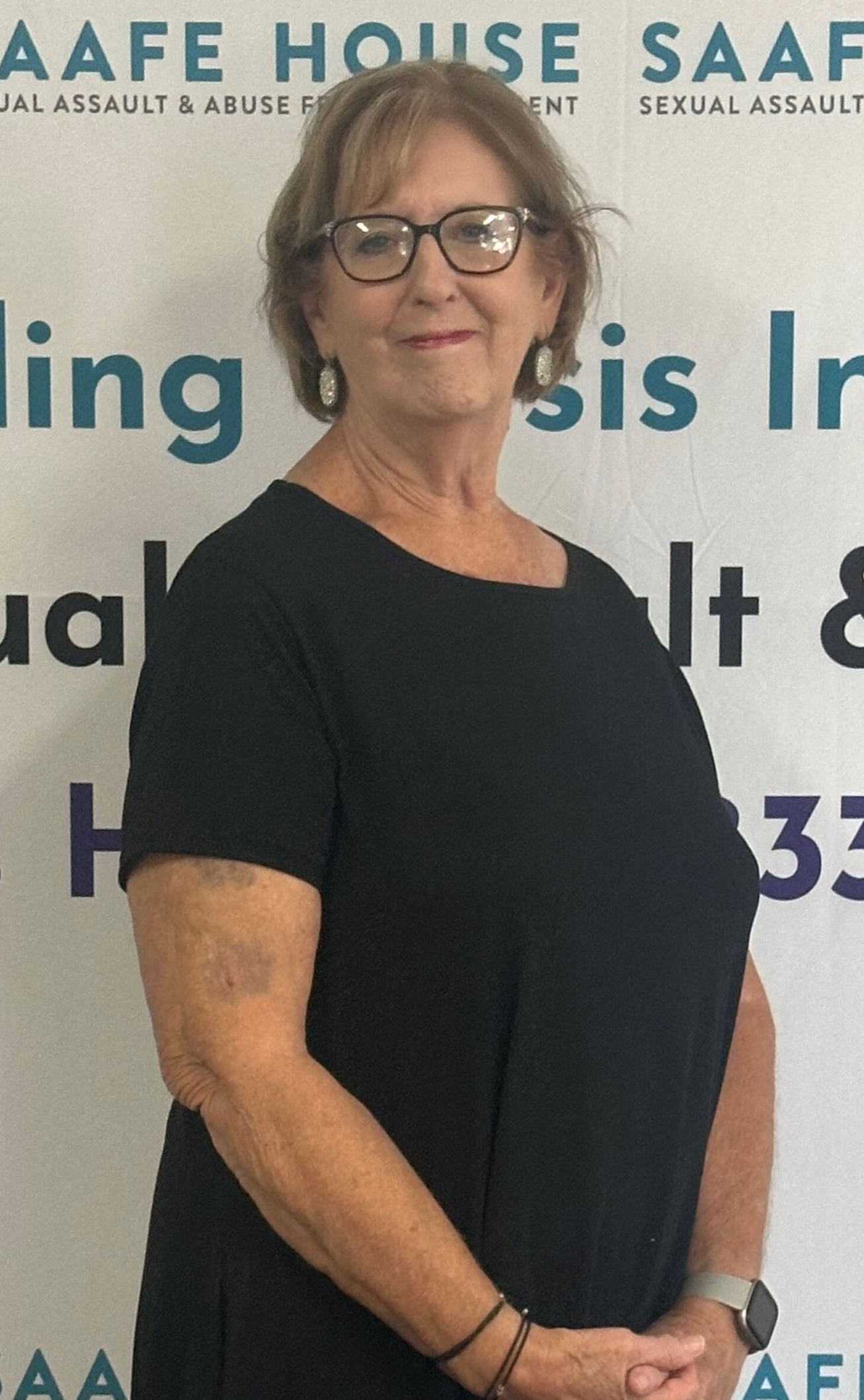 Woman in black shirt and glasses stands in front of a SAAFE HOUSE banner.
