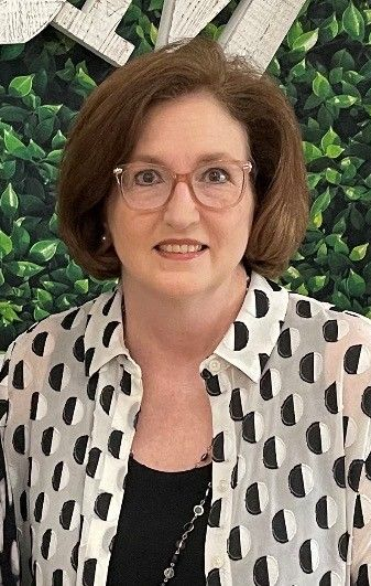 Woman with glasses and short brown hair, wearing a black and white polka dot shirt, smiling in front of a green backdrop.