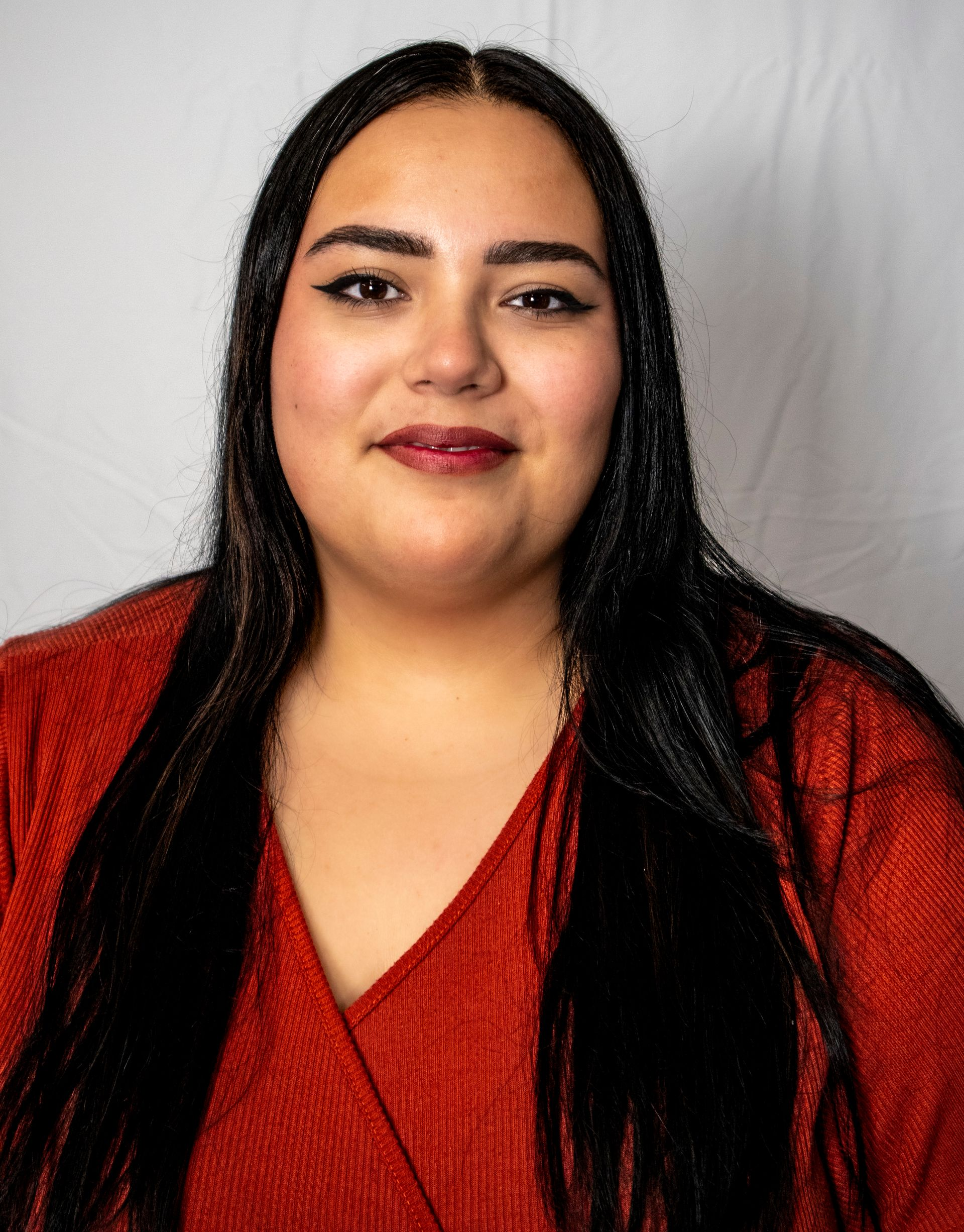 Woman with long, dark hair wearing a red shirt, smiling at the camera against a white backdrop.