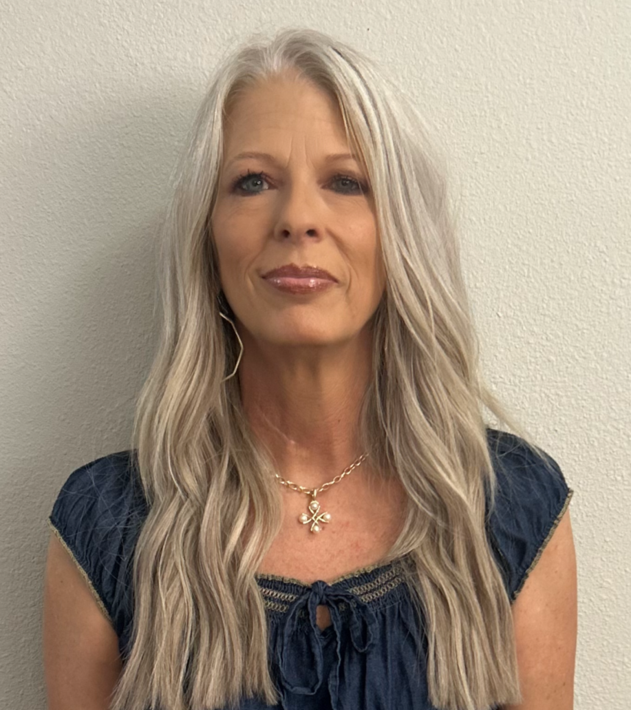 Woman with long gray hair, wearing a blue shirt and necklace, looking at the camera.