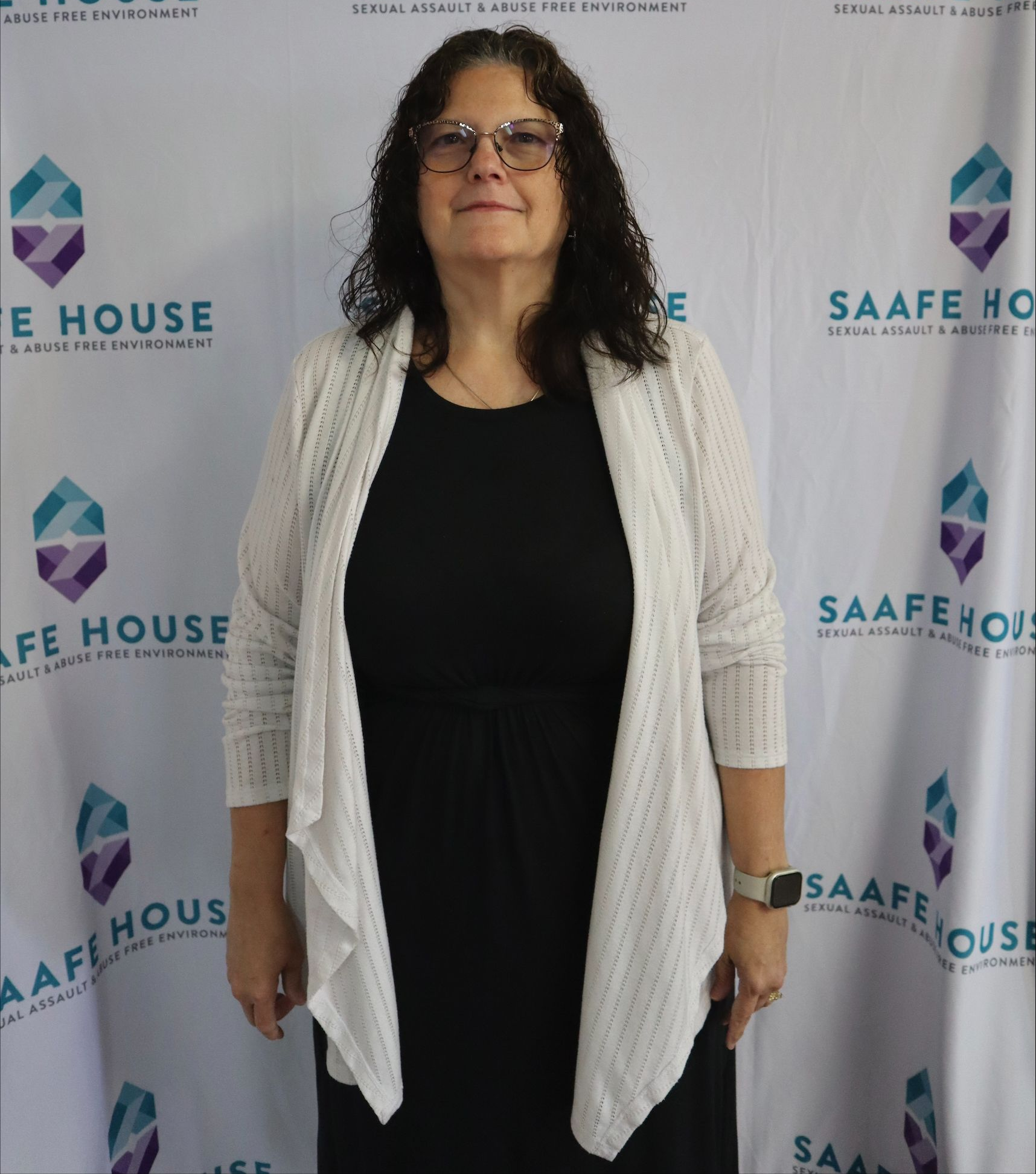 Woman in glasses, black dress, and white cardigan poses in front of a SAAFE HOUSE backdrop.
