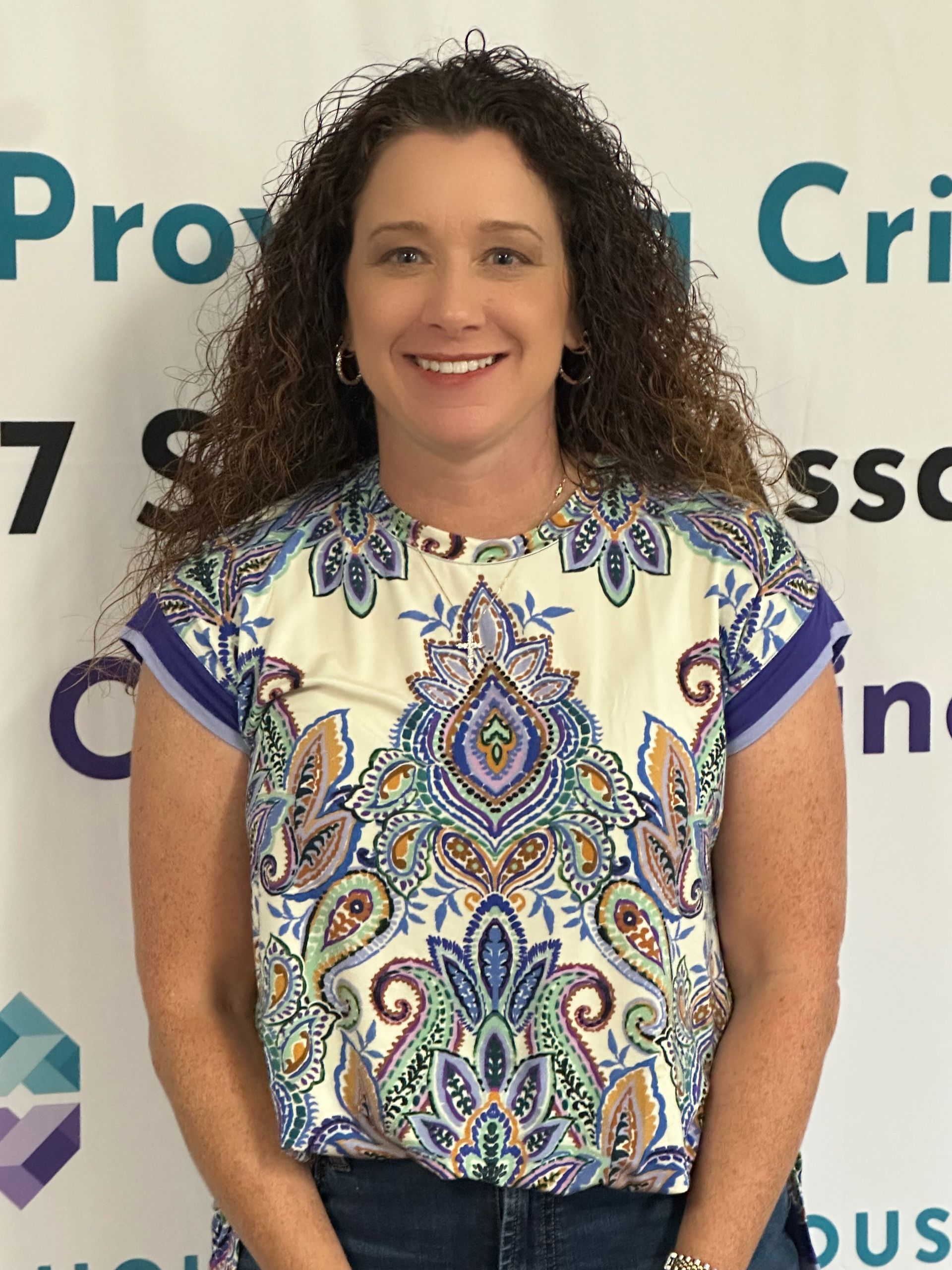 Woman smiling, wearing a paisley print blouse and jeans, standing in front of a sign.