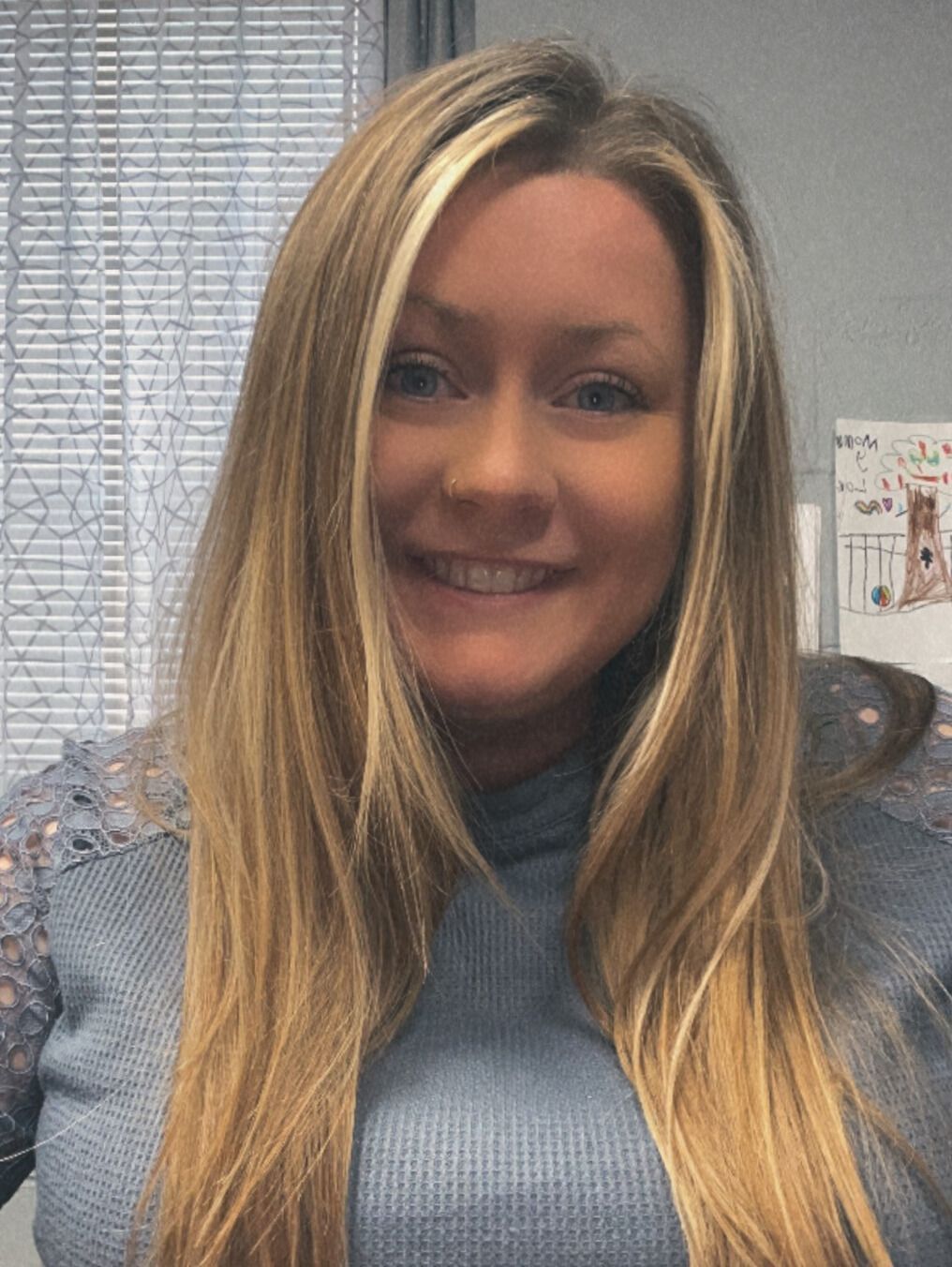 Woman with long blonde hair smiles; she's wearing a blue sweater, indoors near blinds.