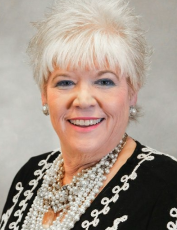 Woman with short, white hair, smiling, wearing pearl necklace and black top with white trim.