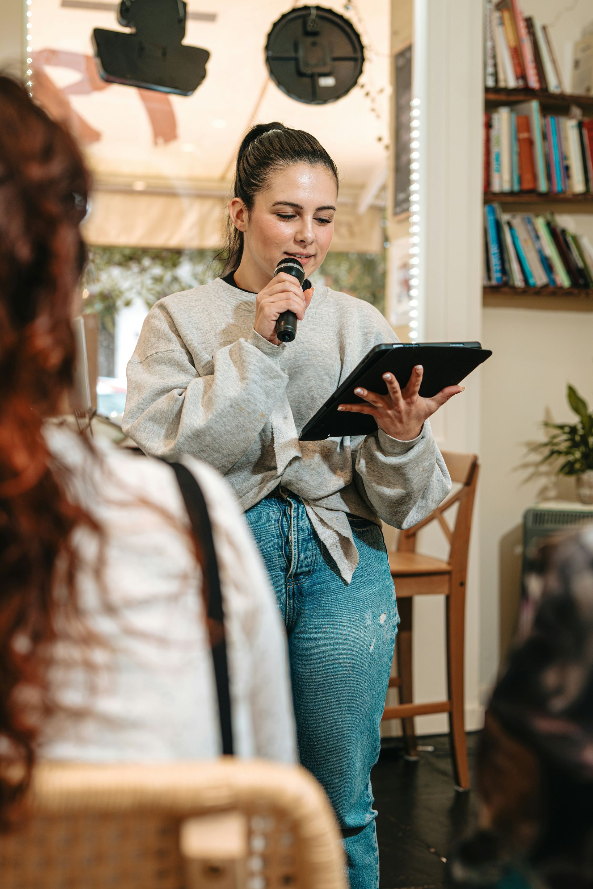 Woman speaking into a microphone, holding a tablet; indoors, light-colored sweater, reading to an audience.