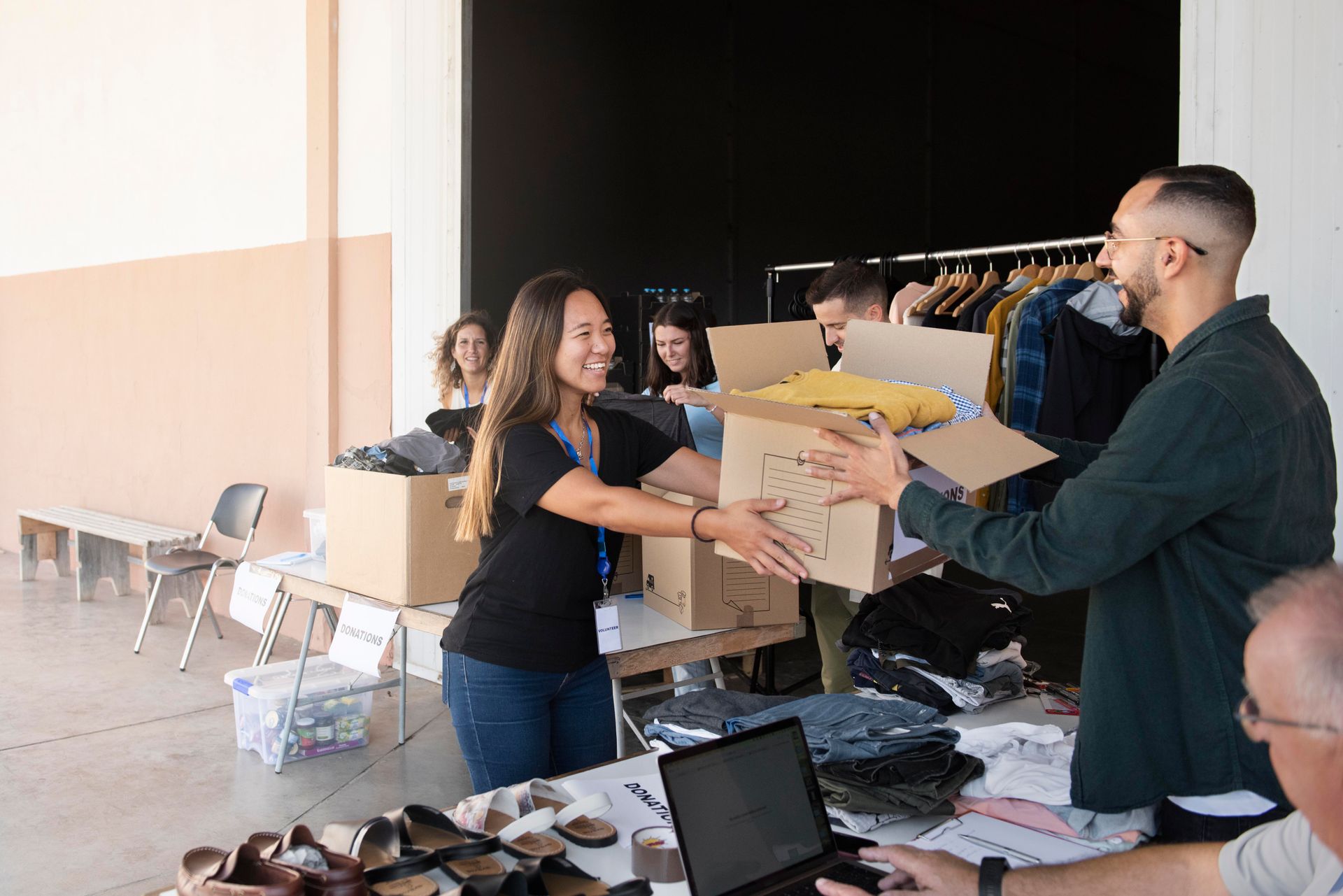 Volunteers sorting donations in a brightly lit room. A woman passes a box to a man, clothes on a rack in the background.