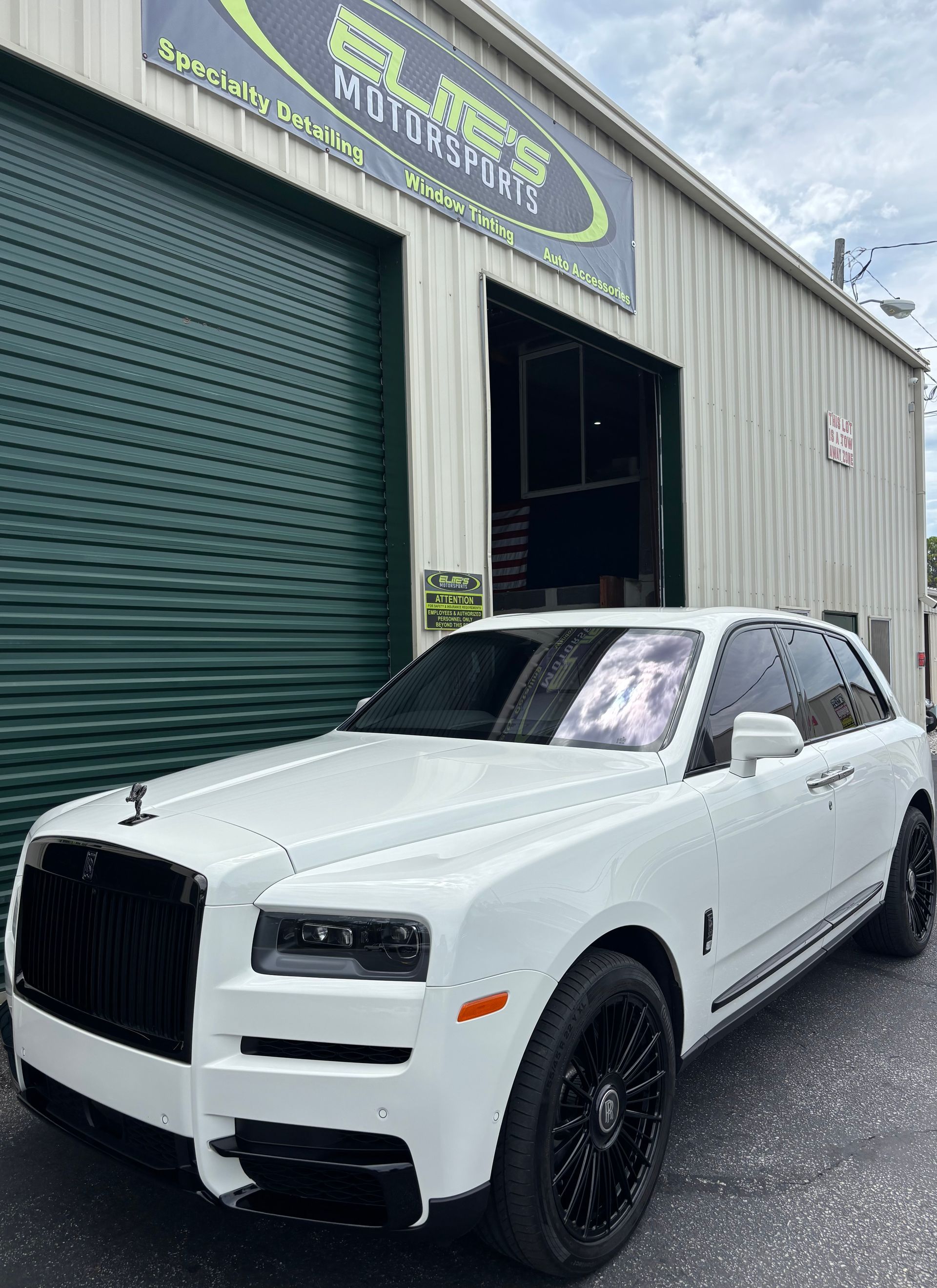 White Rolls-Royce SUV parked in front of a garage, black wheels and trim, under a cloudy sky.