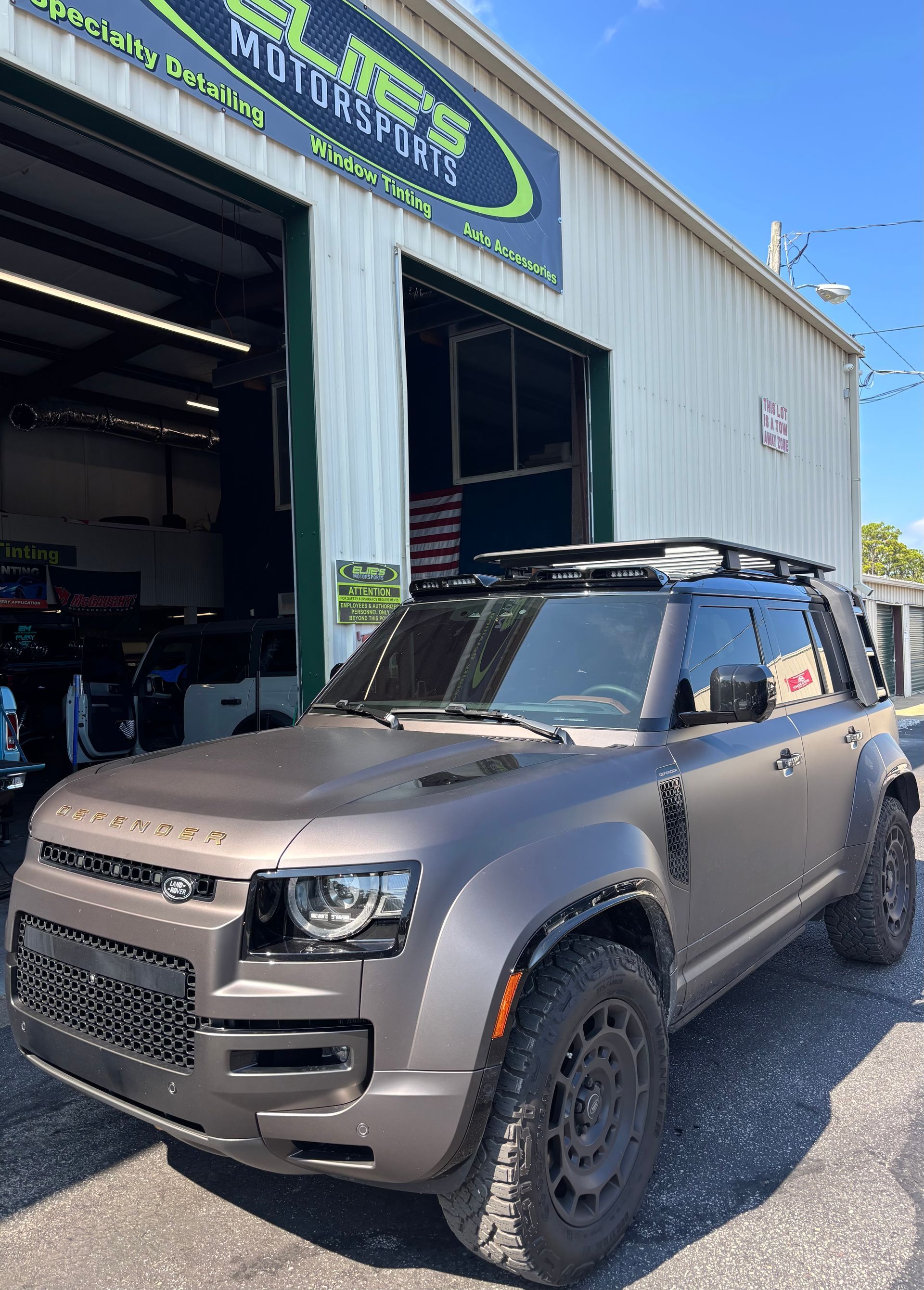 Brown Land Rover Defender SUV outside a motorsports shop with black wheels and a roof rack.