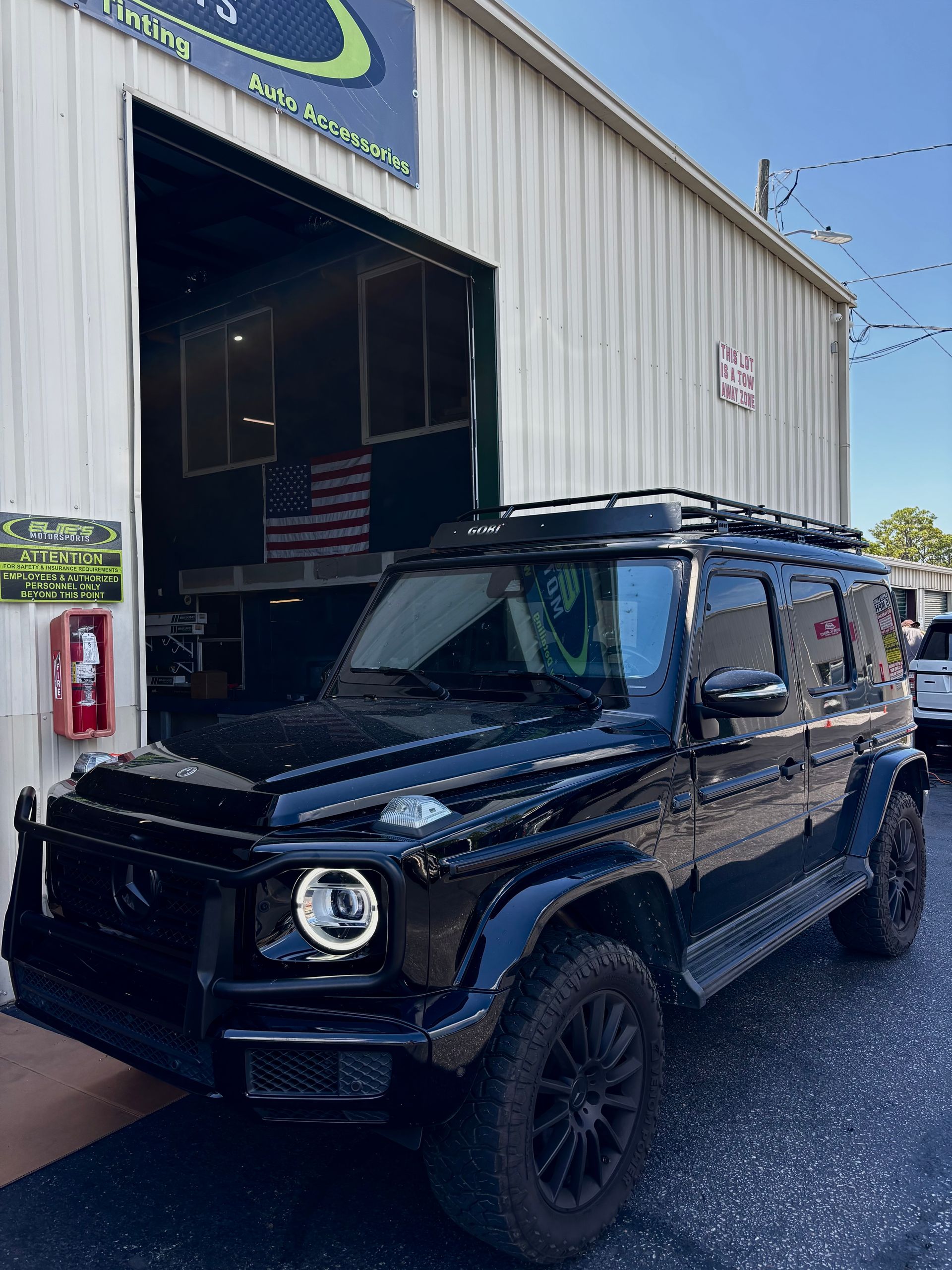 Black Mercedes-Benz G-Wagon parked in front of a garage, with a roof rack and brush guard.