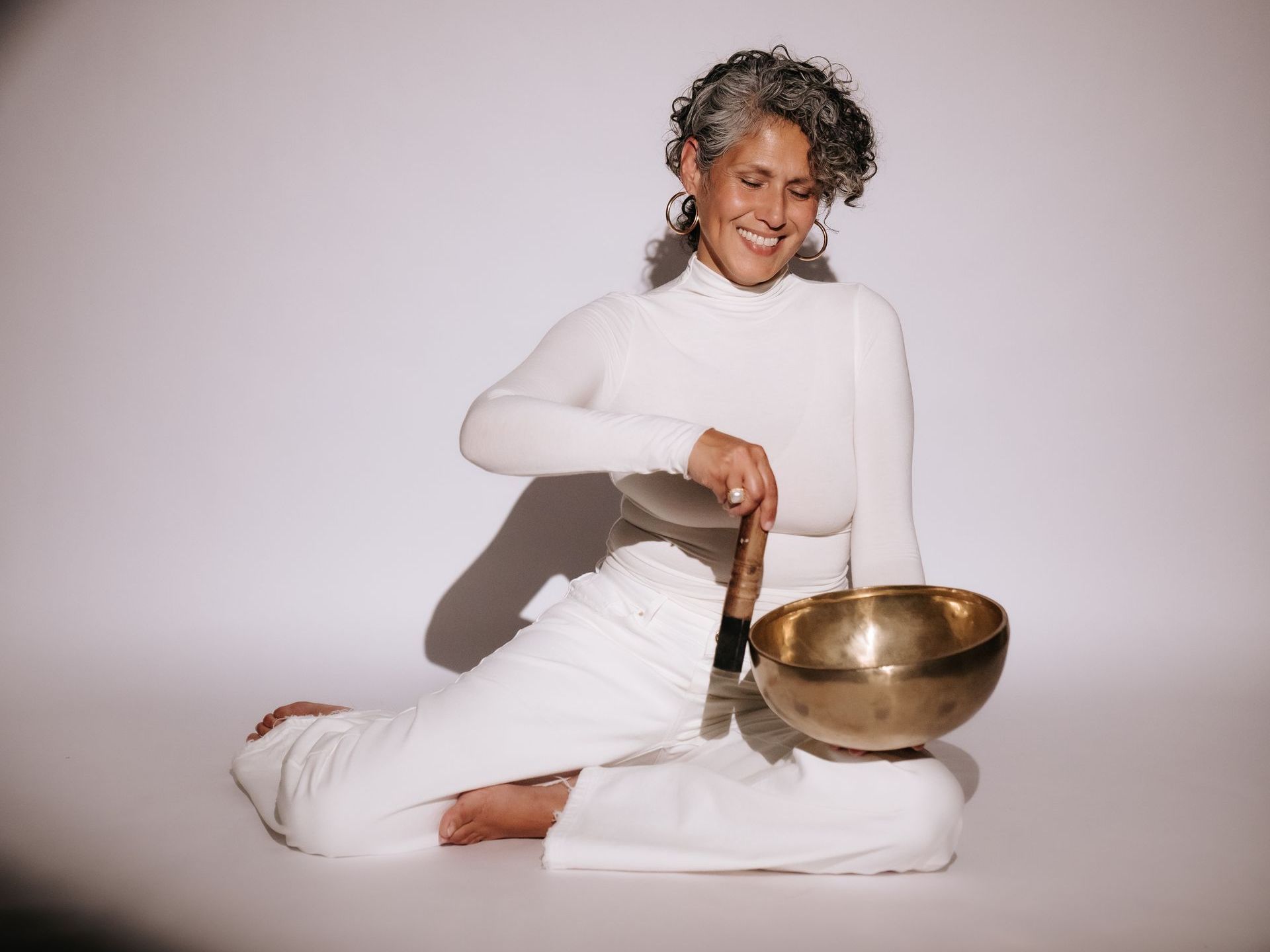 Vrouw in het wit die glimlachend op de grond zit en een gouden klankschaal bespeelt – symbool voor licht, klank en heling.