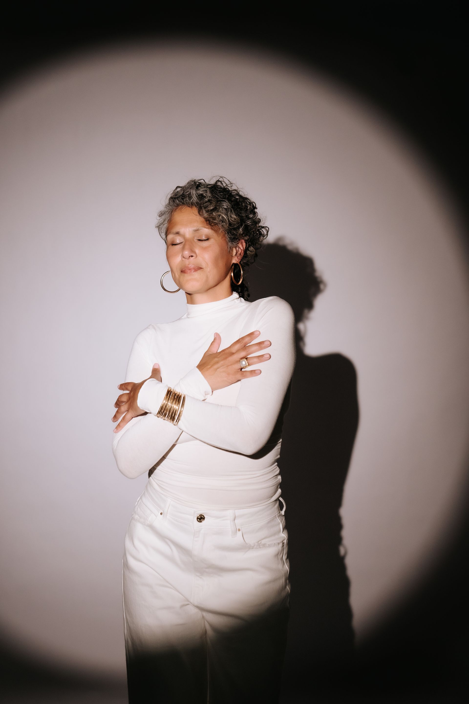 Woman in white clothing hugs herself, eyes closed, against a light-washed wall.