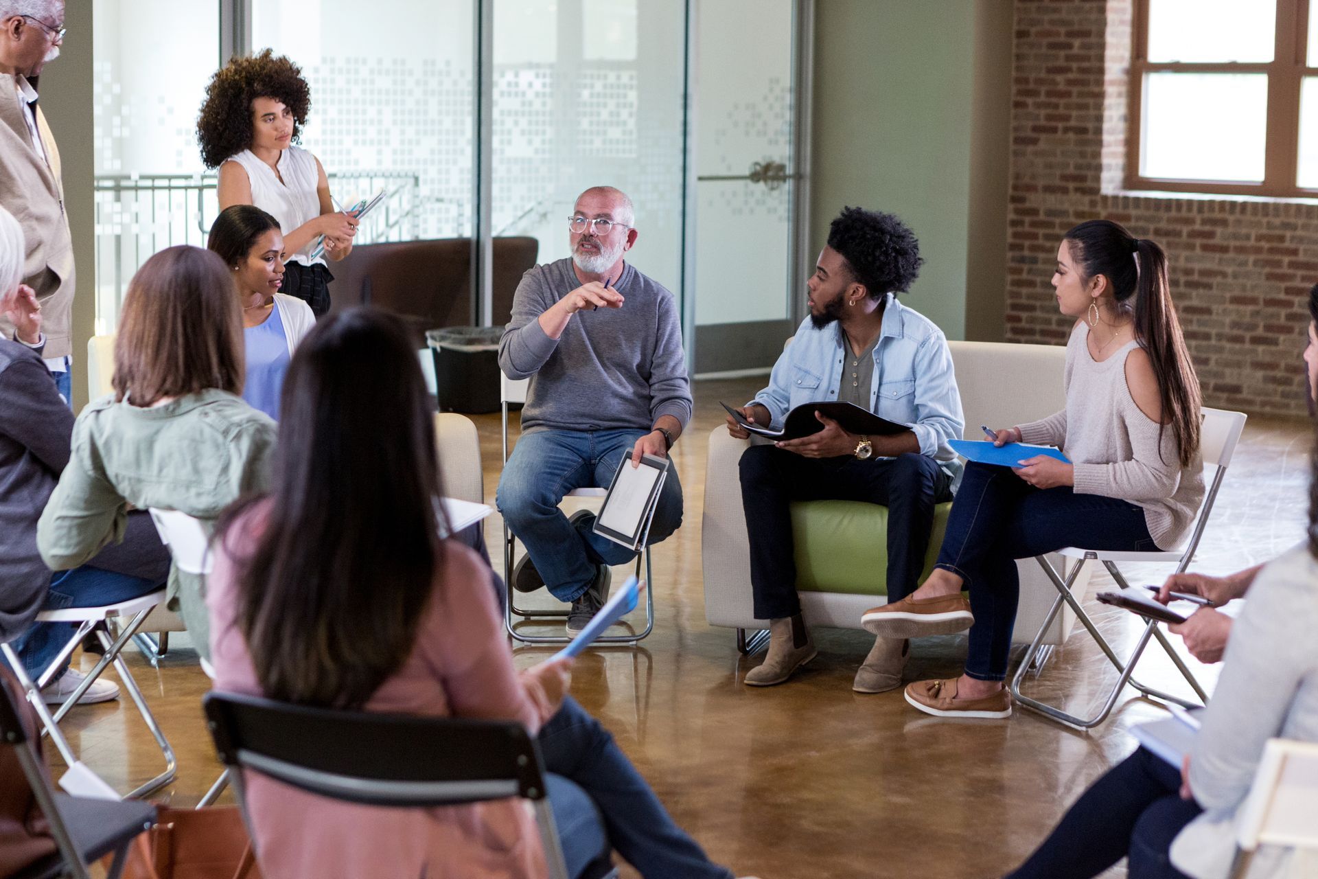 Group of people in chairs, discussing in a circle. A man gestures while speaking; others listen.