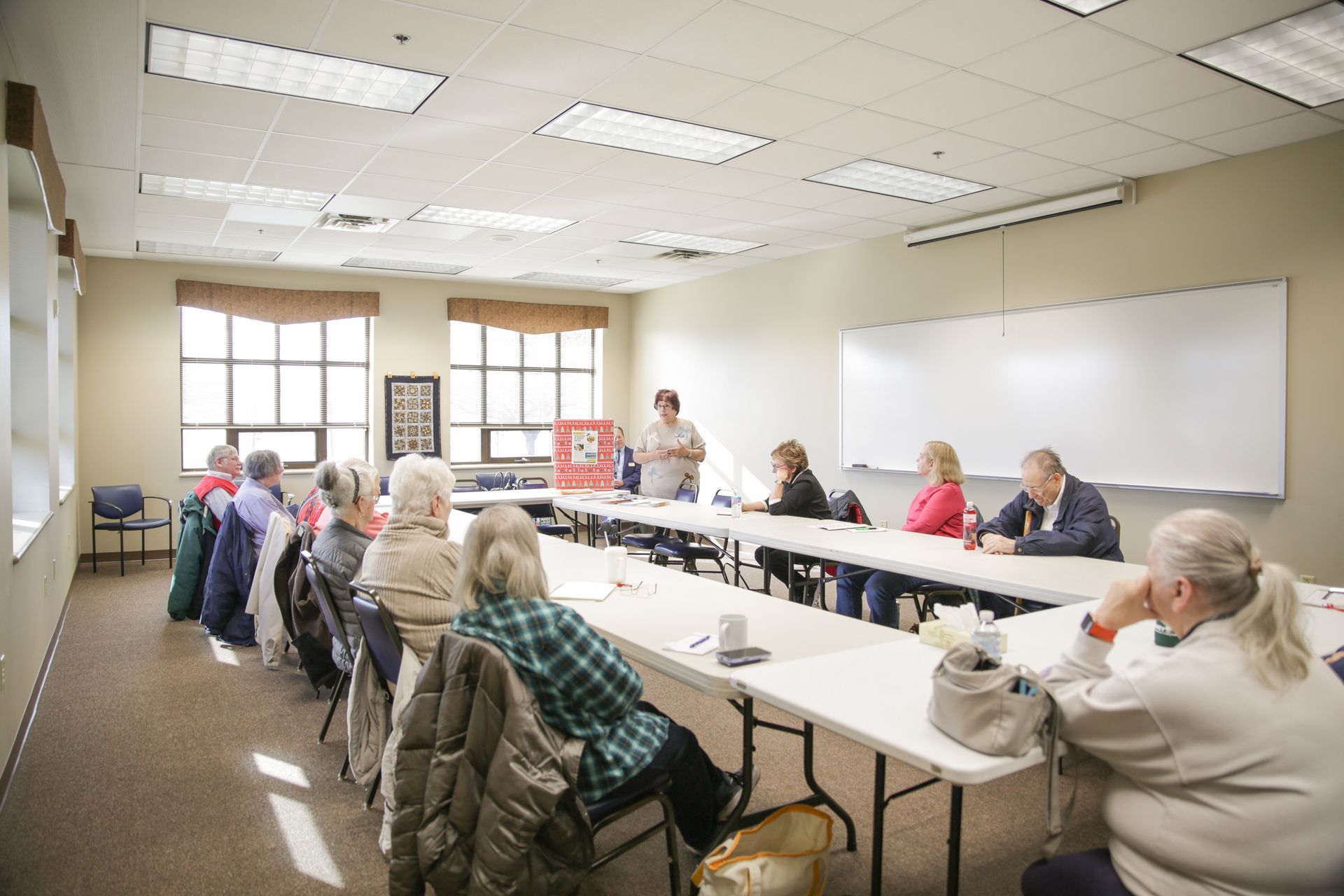 Group of people seated at a long table in a brightly lit room, listening to a speaker.