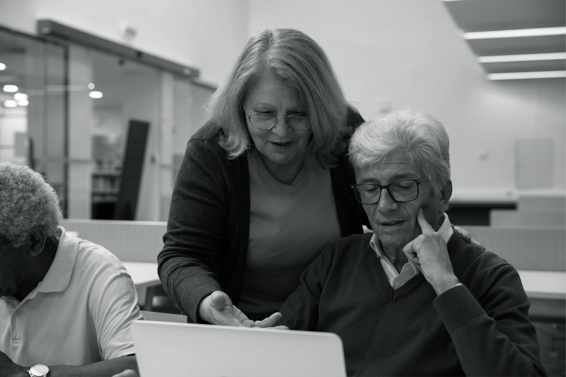 Elder woman helping an elder man at a computer. They're both wearing glasses and looking at the screen. A third person is seated nearby.