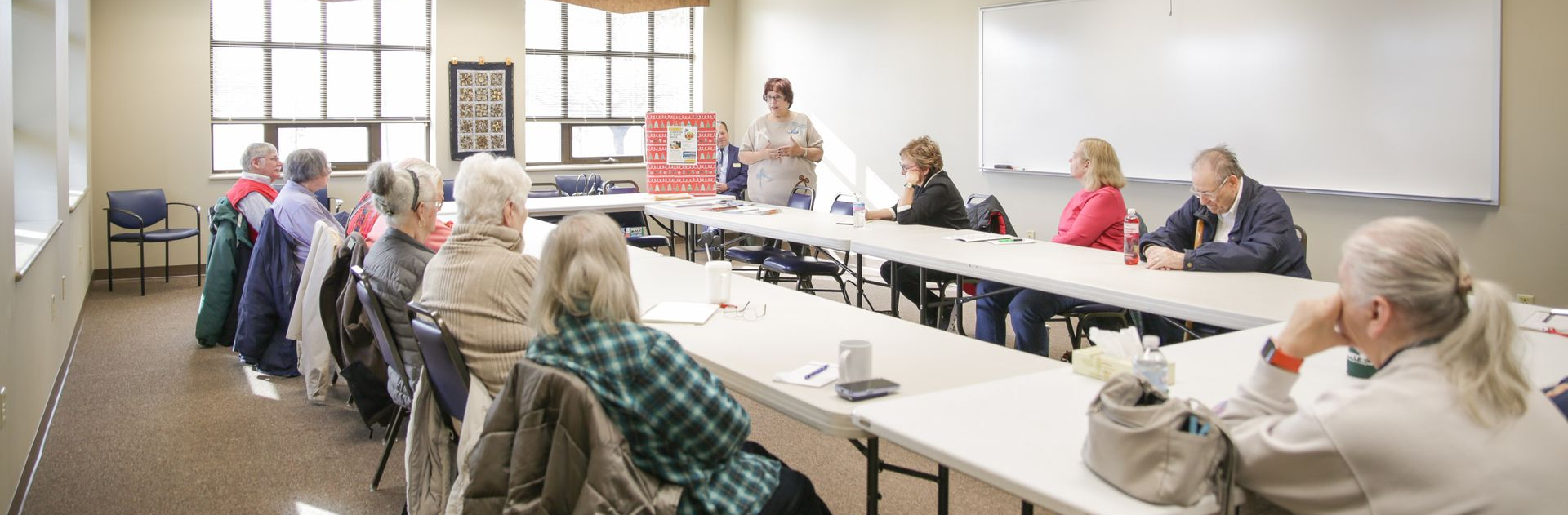A group of people are seated at a long table in a room with a presenter at the front.