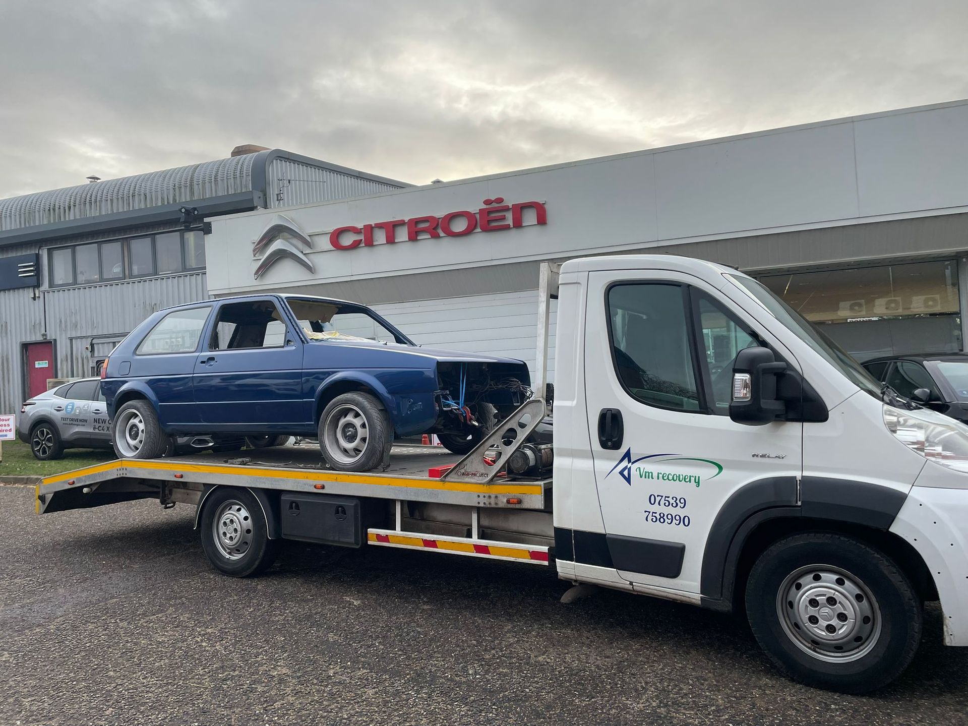 A tow truck with a blue car on the back is parked in front of a citroen dealership.