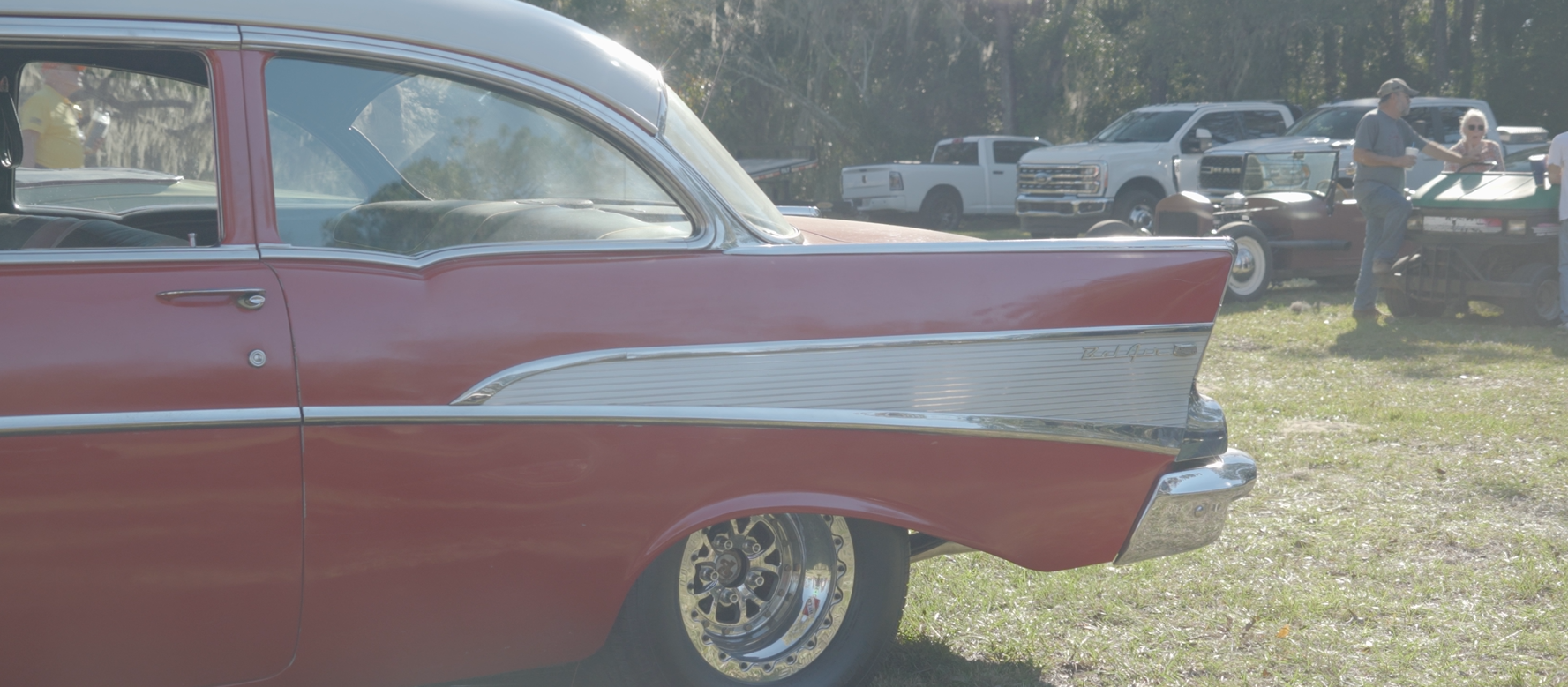 A classic red car with a white top, parked on grass. Other vehicles and people are in the background.