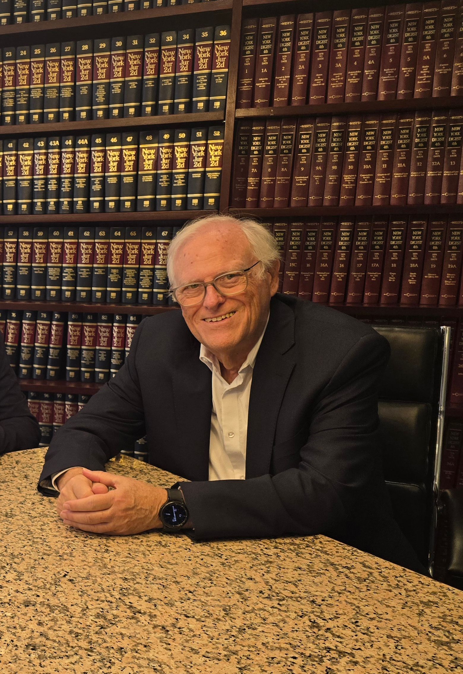 Man in glasses, sitting at a table in a library with law books, smiling.