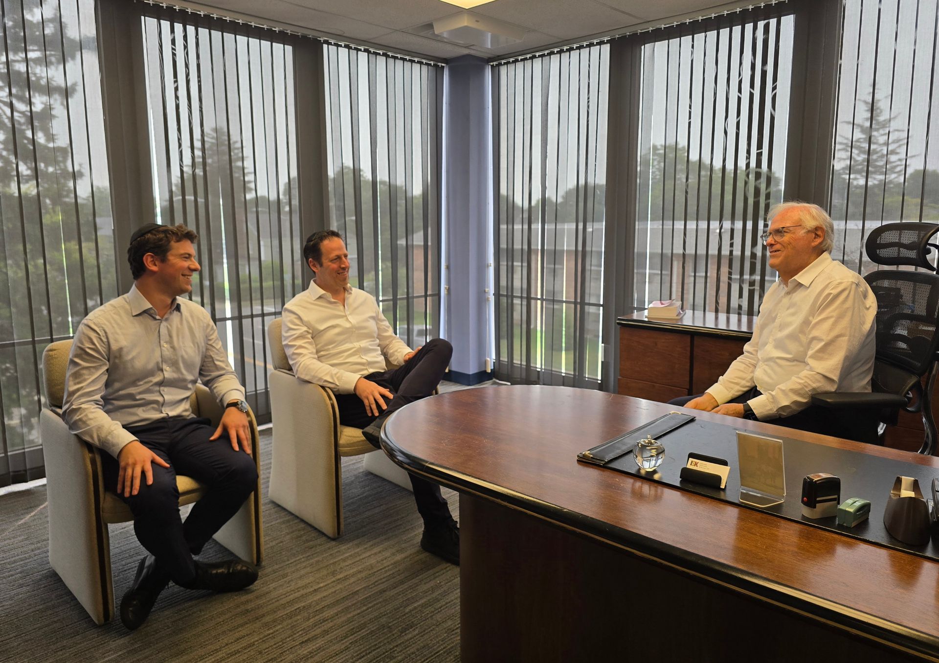 Three men in a bright office; two seated smiling, the other at a desk. Light floods the room through window blinds.