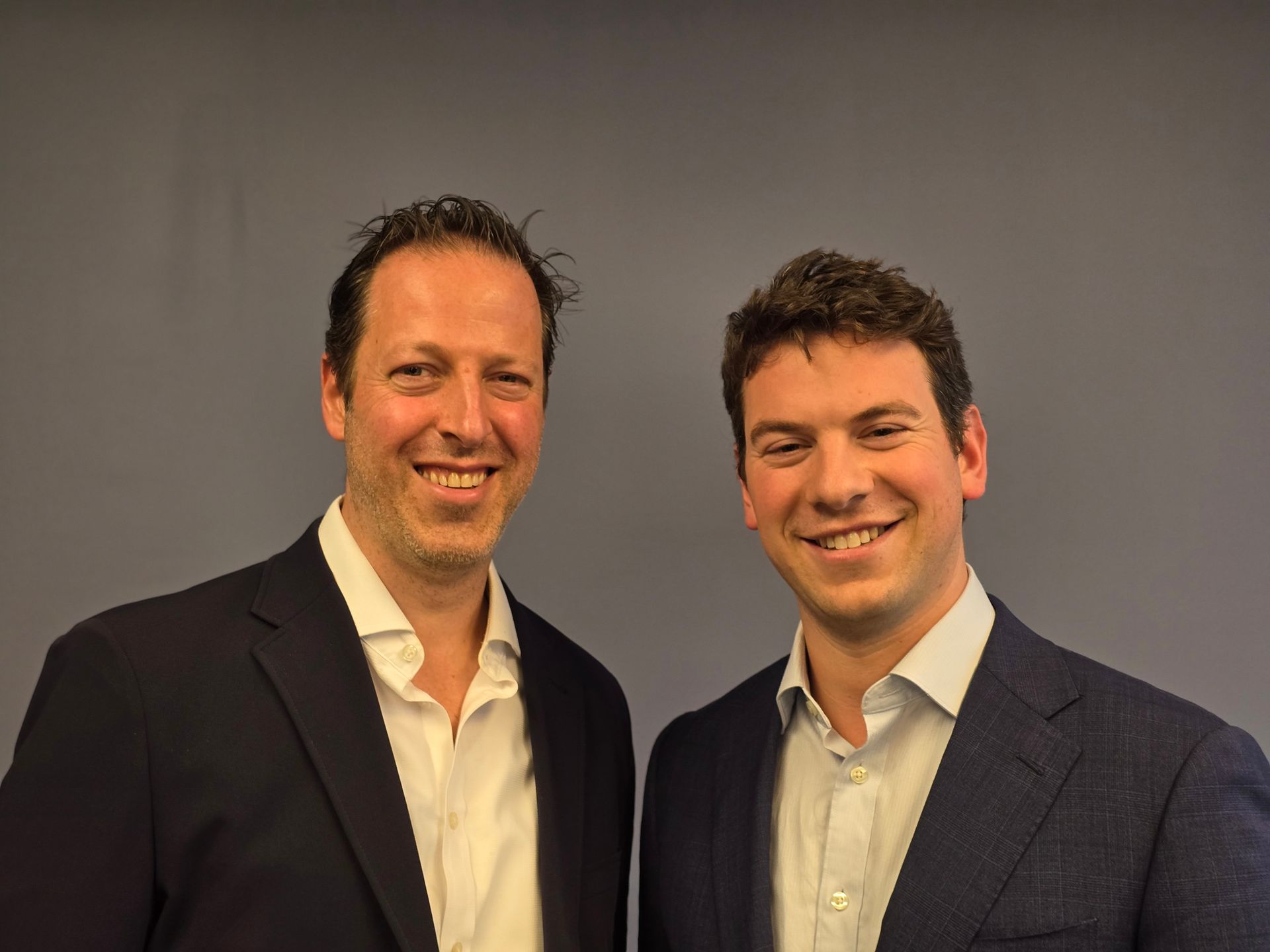 Two men smiling, wearing suits, posing in front of a gray wall.