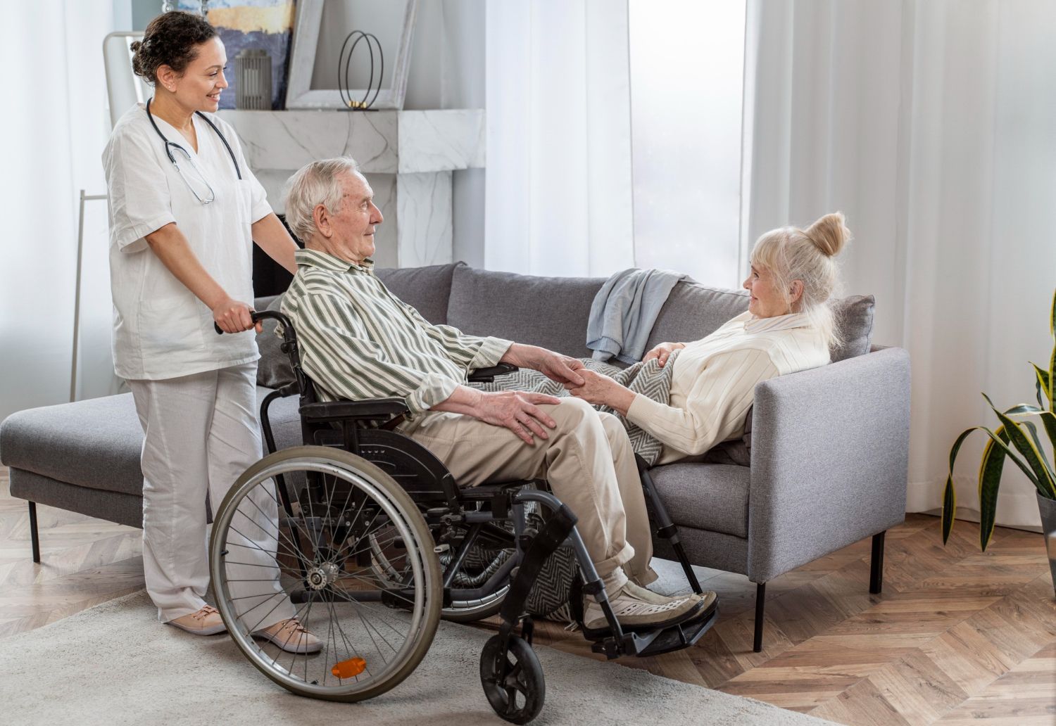 Caregiver assisting an older man in a wheelchair; a woman lies on the sofa, holding his hand. Living room setting.