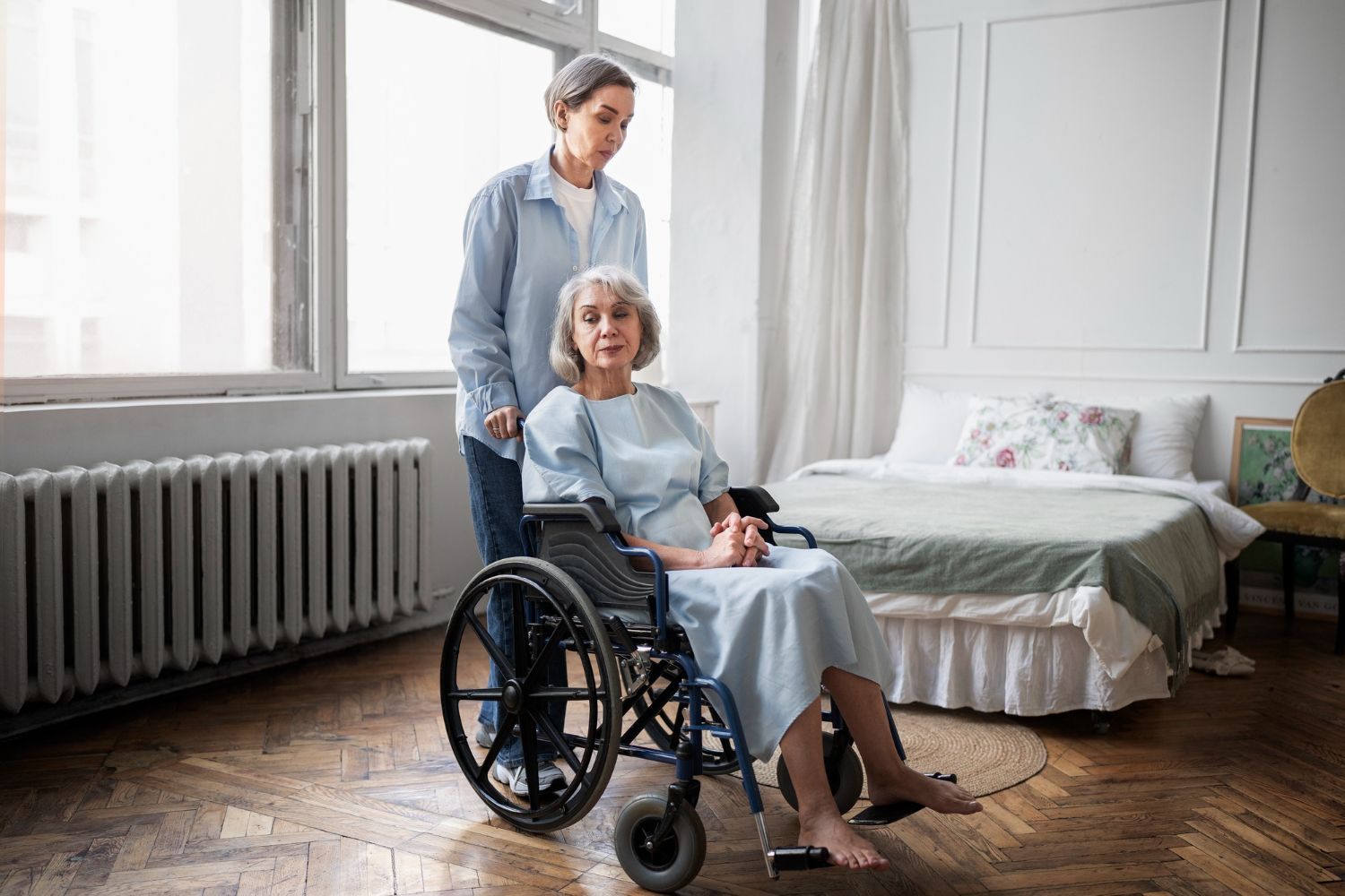 Woman in wheelchair, assisted by another woman, in a bedroom with large window and bed.