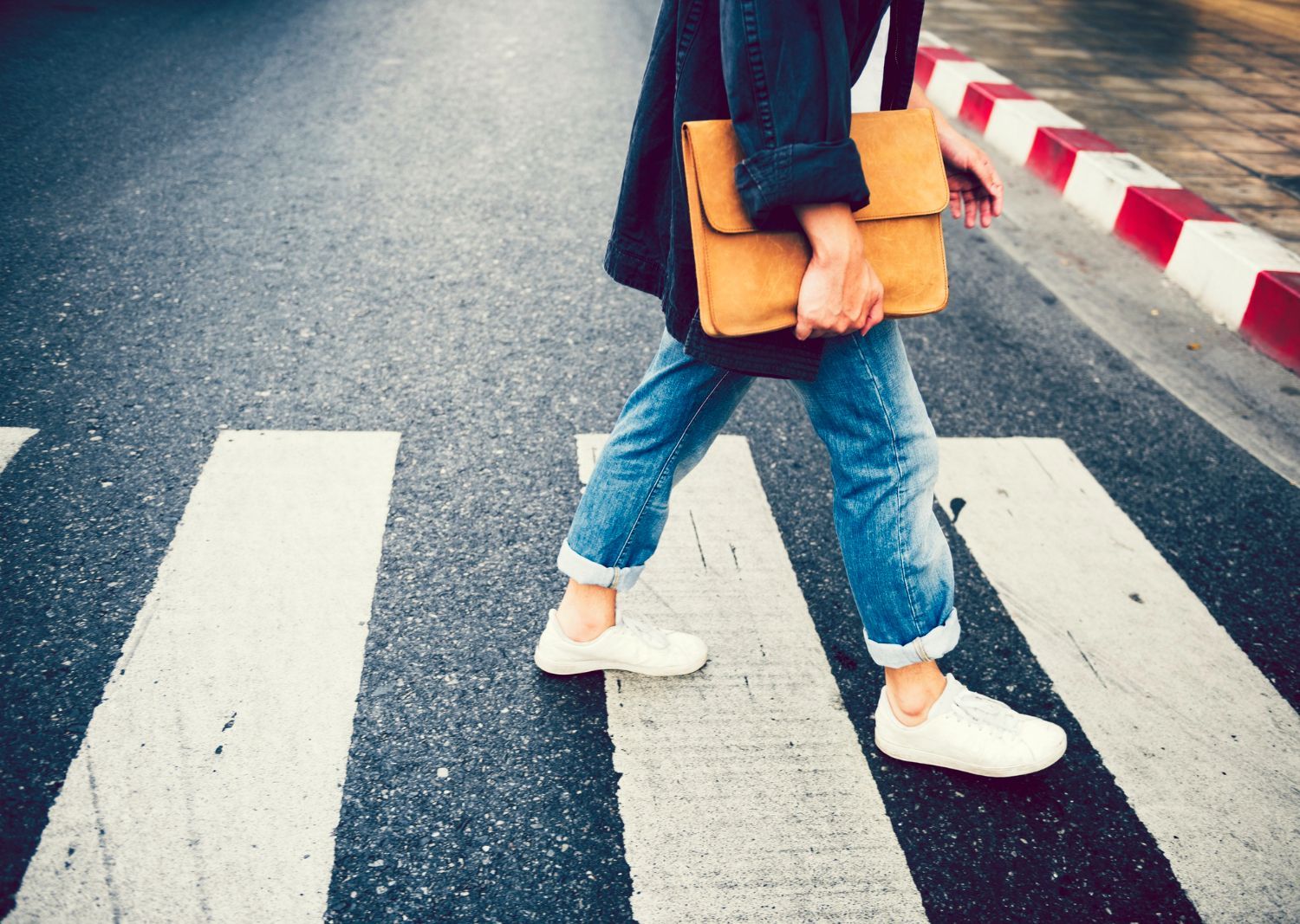 Person walking across a crosswalk, holding a tan bag. White sneakers, blue jeans, and dark jacket.