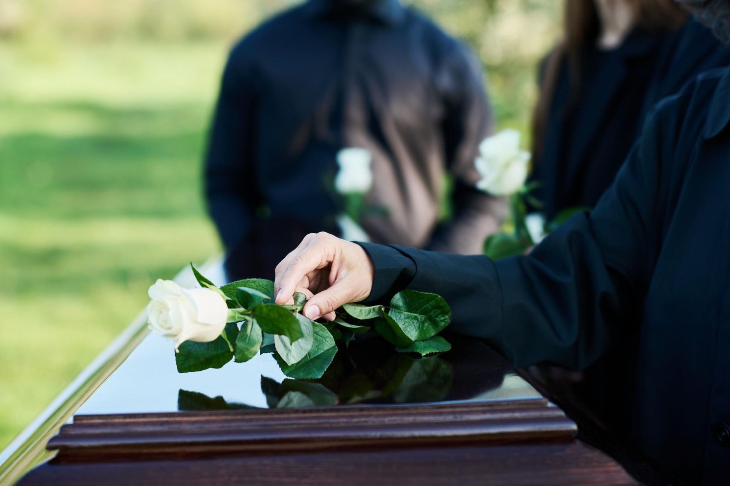 Hand placing a white rose on a closed casket at a funeral. People in black attire stand nearby.