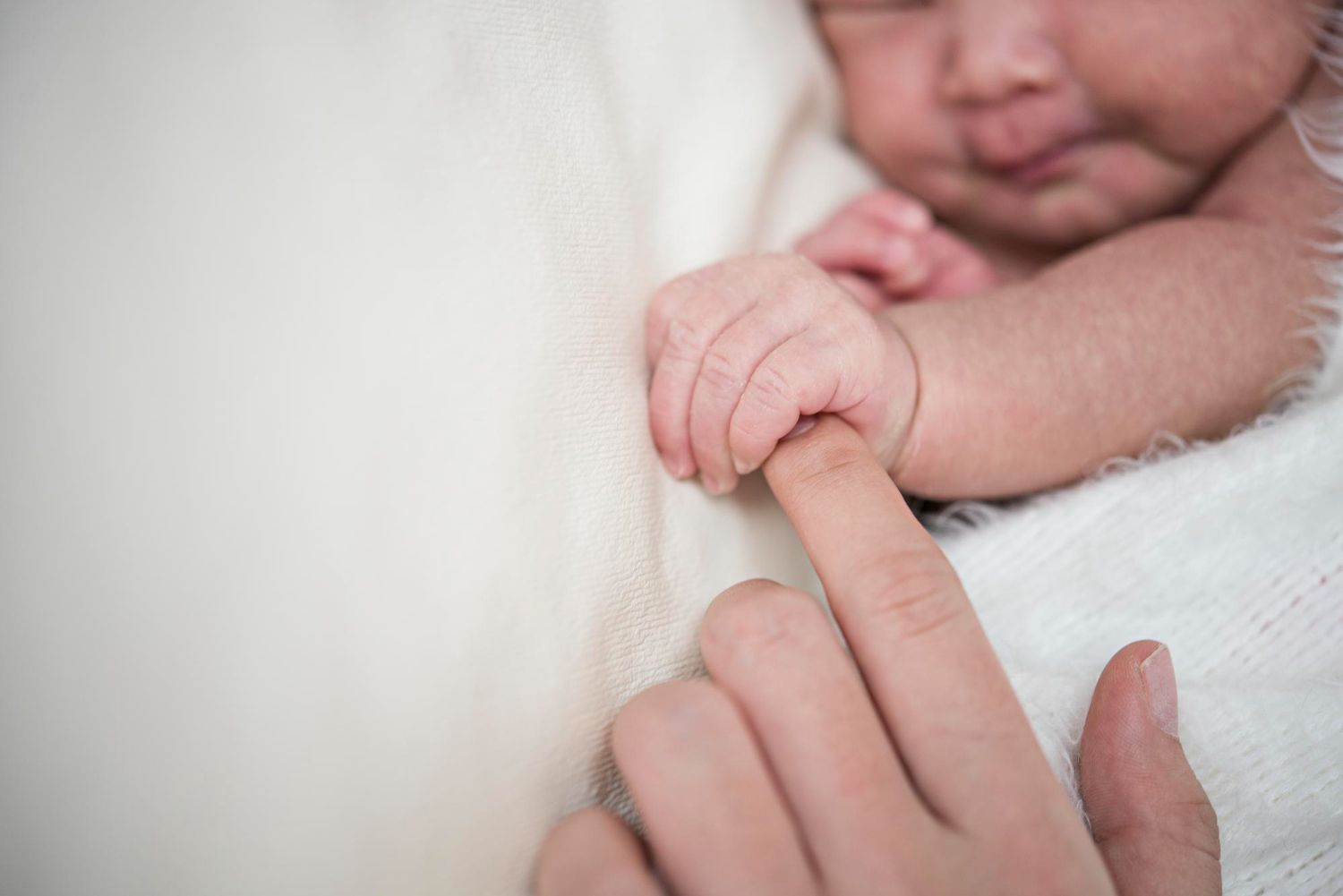 Baby's hand holding an adult finger, close-up, soft focus, white blanket.